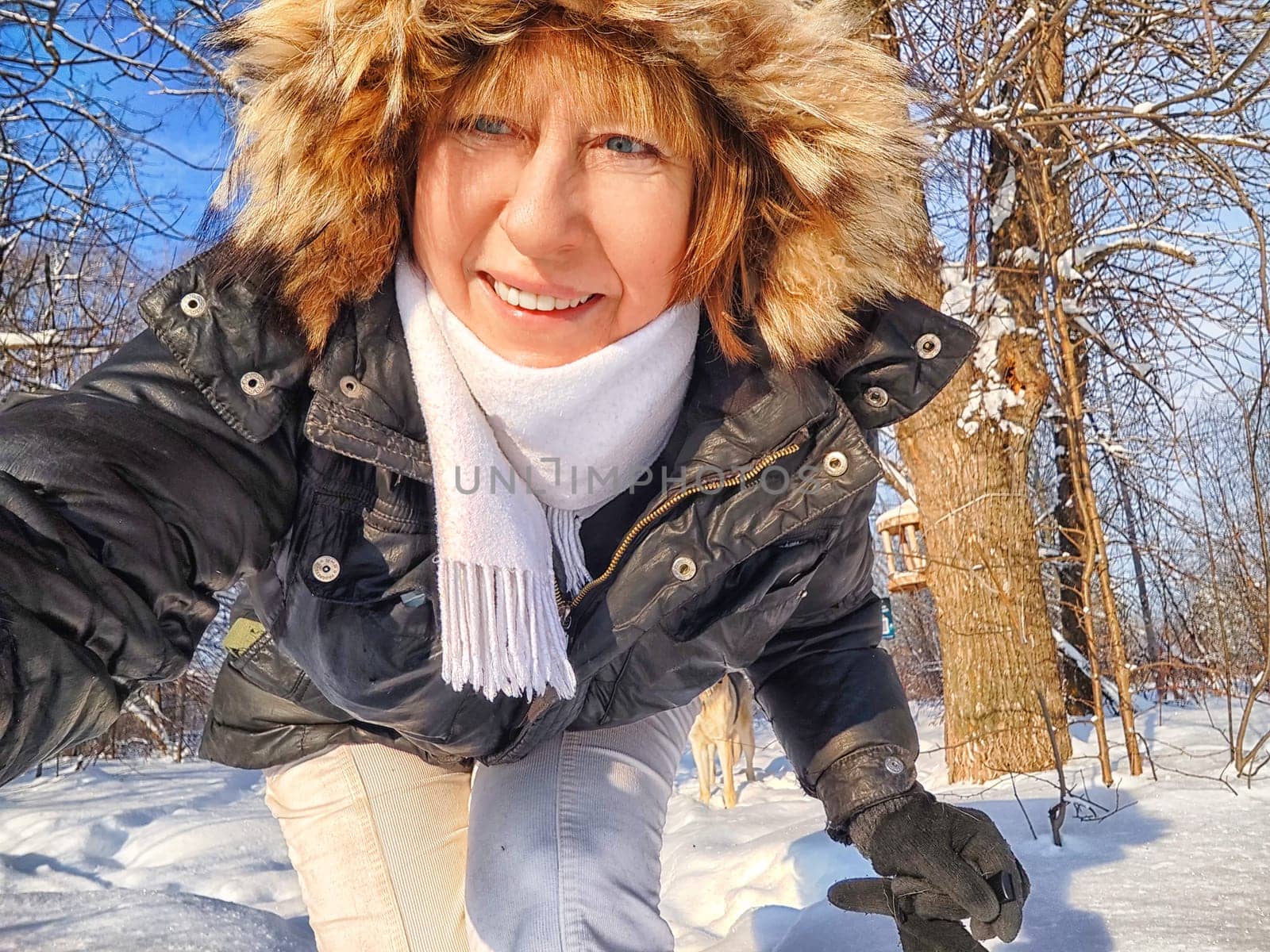 A cheerful middle aged woman in a winter coat with fur, scarf taking selfie on nature outdoors and cold, snowy background with blue sky and white clouds