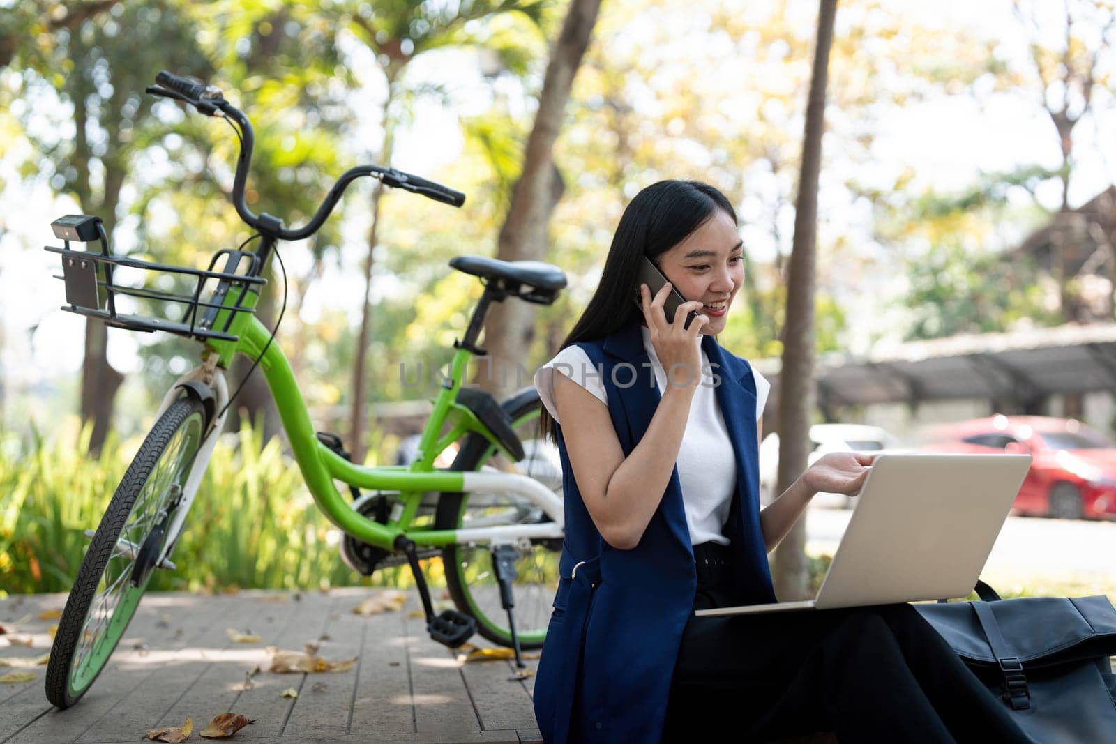 Asian businesswoman with bicycle using laptop and smartphone and sitting outside the office building. Woman commuting on bike go to work. Eco friendly vehicle, sustainable lifestyle concept by nateemee