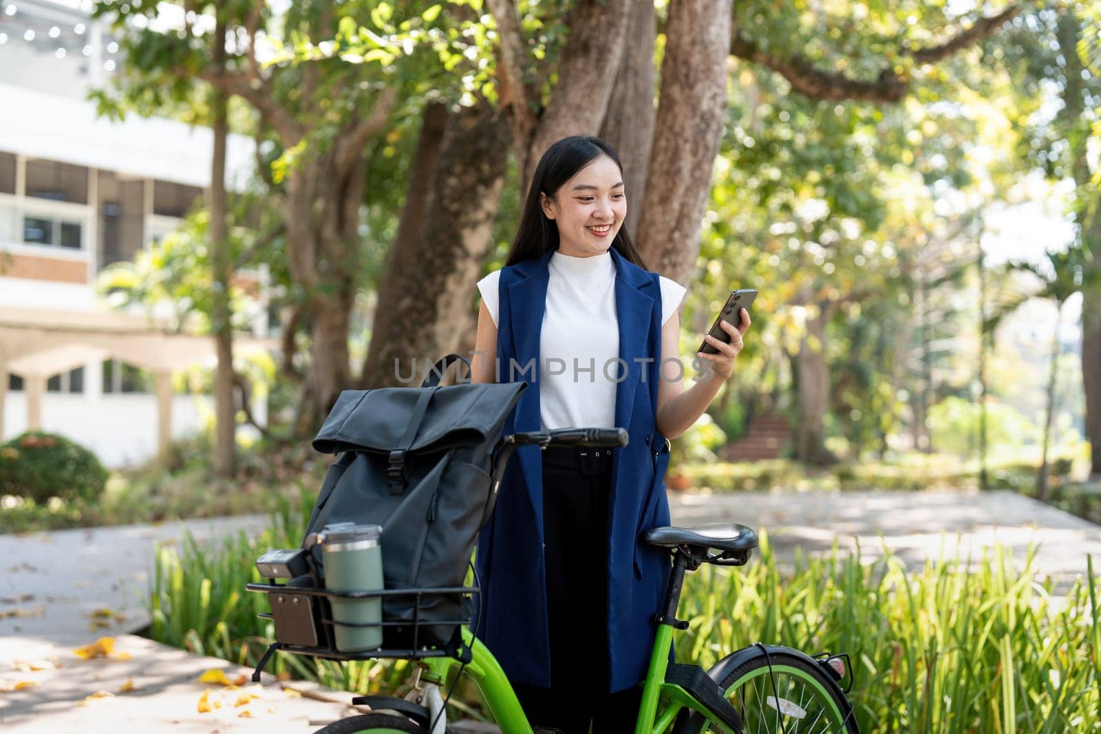Asian businesswoman with bicycle using smartphone and sitting outside the office building. Woman commuting on bike go to work. Eco friendly vehicle, sustainable lifestyle concept.