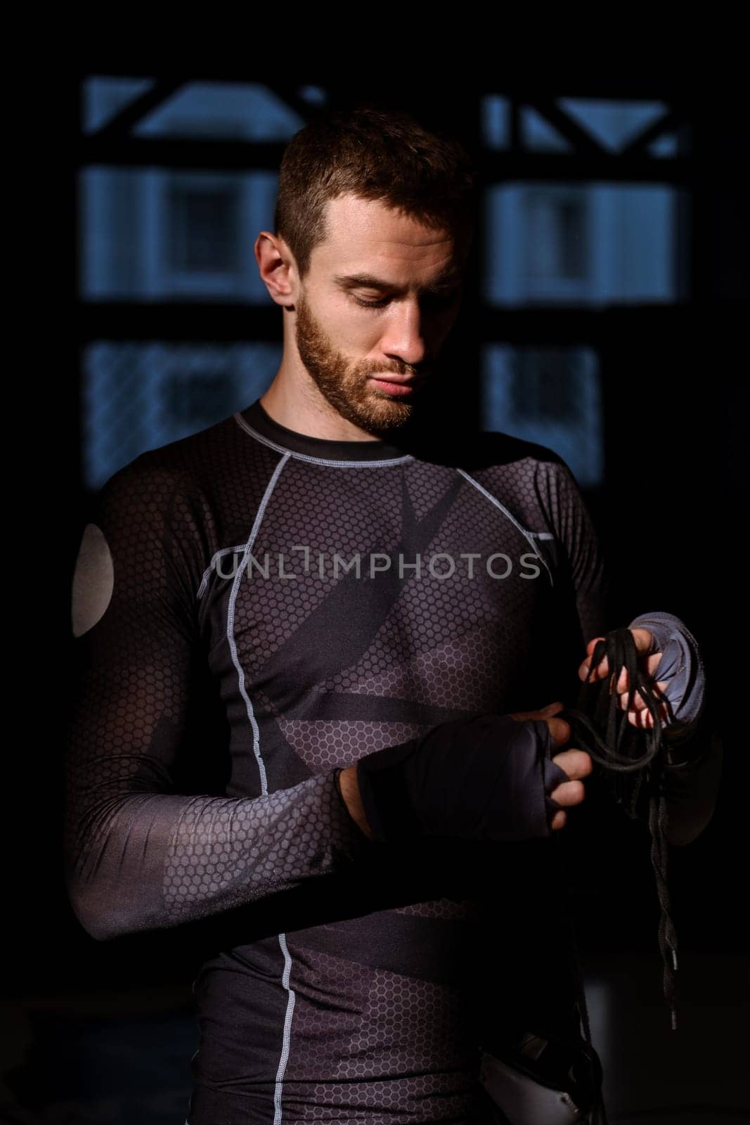 Confident composed young bearded man in sportswear, with wrists wrapped in bandages, standing in rays of sunlight in dark gym holding boxing gloves, preparing for training