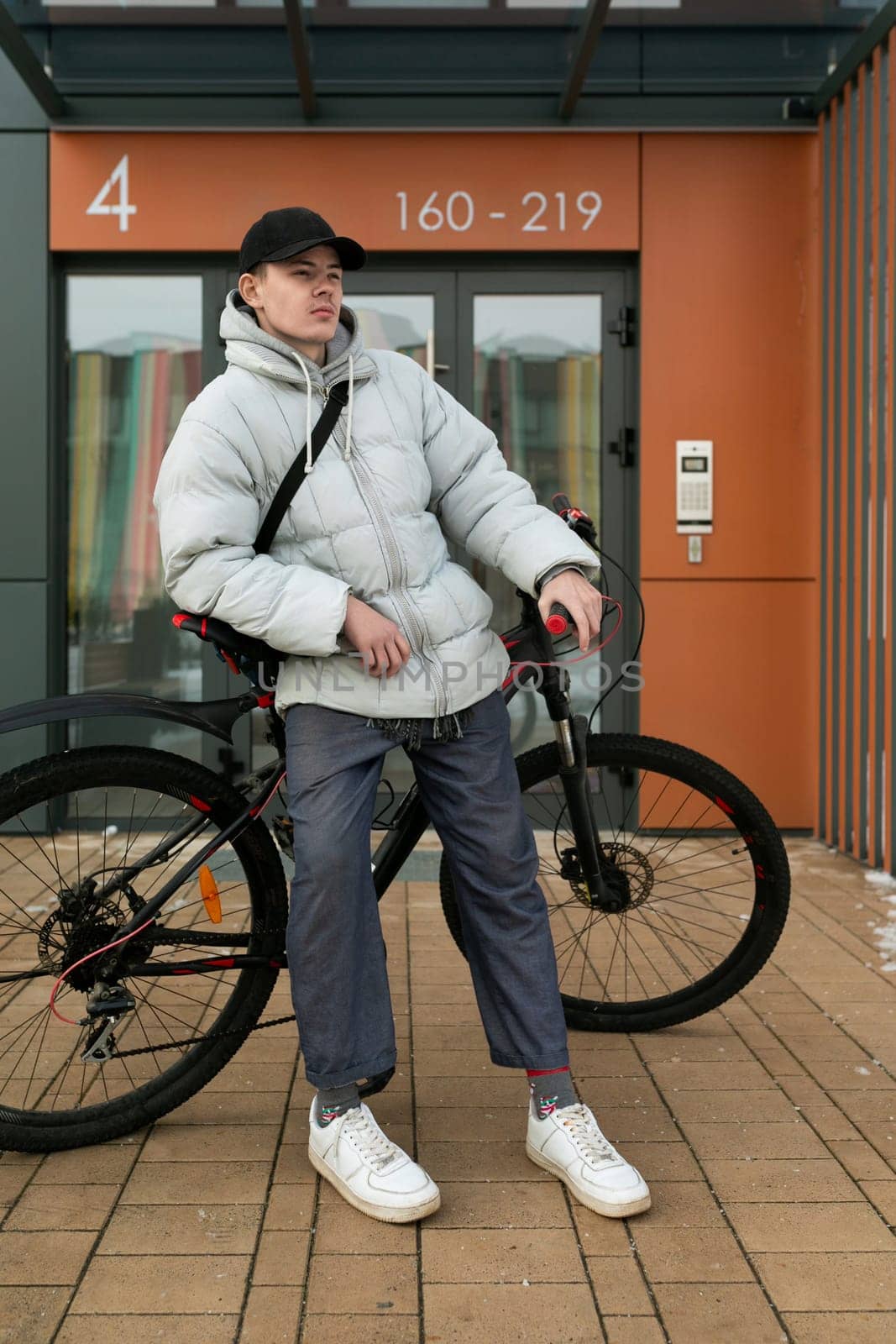 A young Caucasian man with a gray jacket and black cap is waiting for a friend with a bicycle under the porch by TRMK
