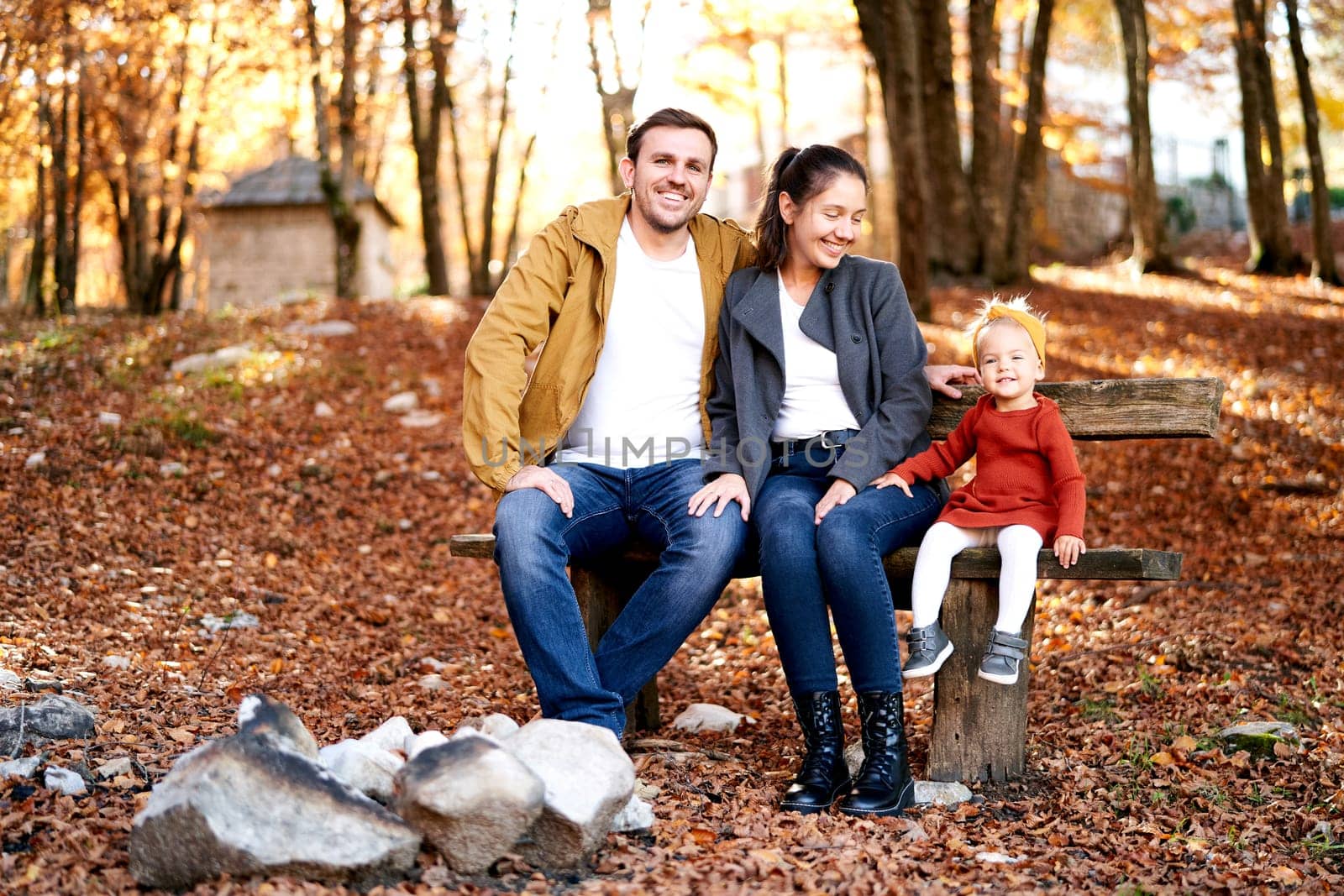 Smiling mom and dad with a little girl are sitting on a bench in the autumn forest. High quality photo