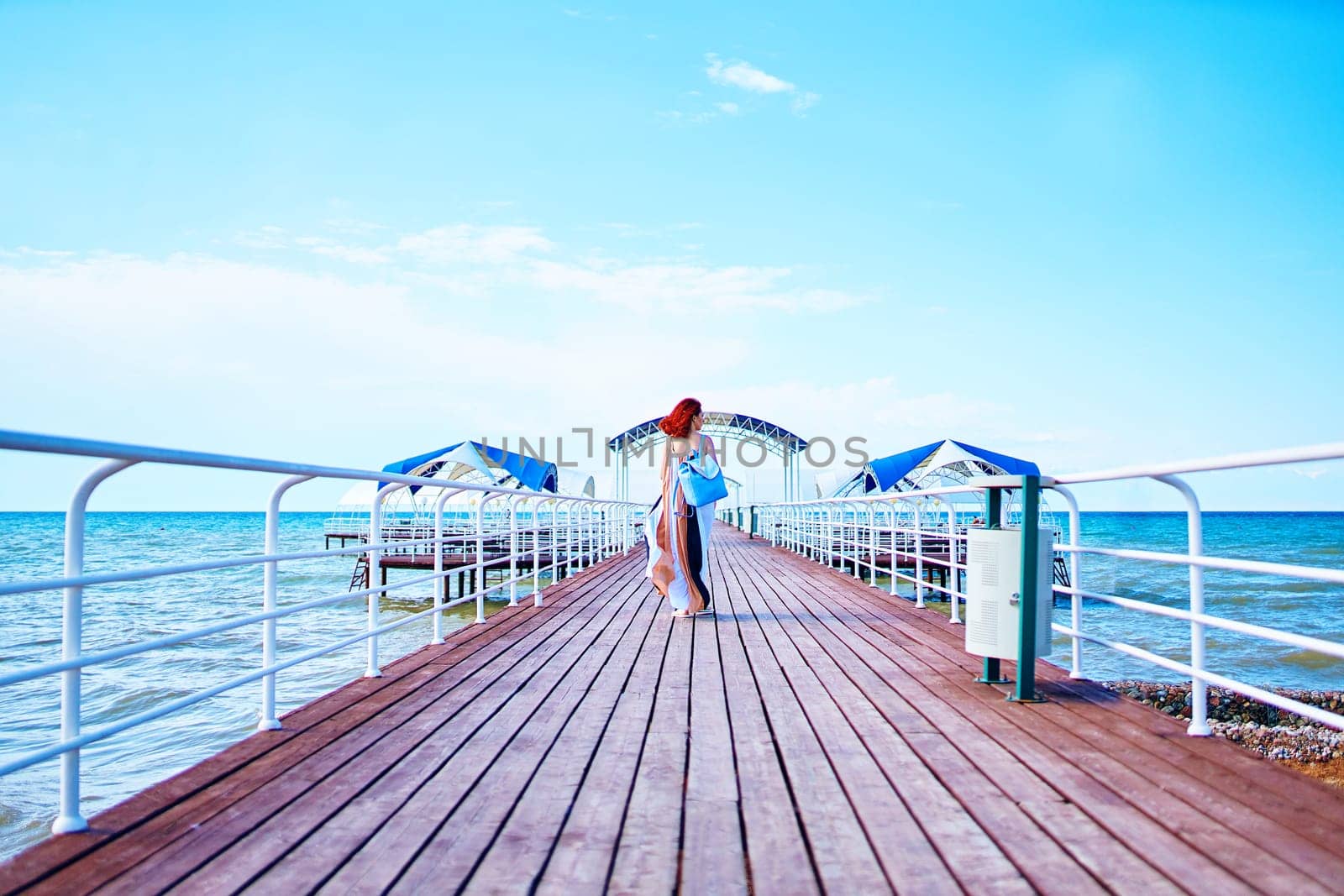 Amidst the resort pier and the tranquil sea, a woman with beautiful red hair, dressed in a summer ensemble and carrying a backpack, takes a leisurely stroll.