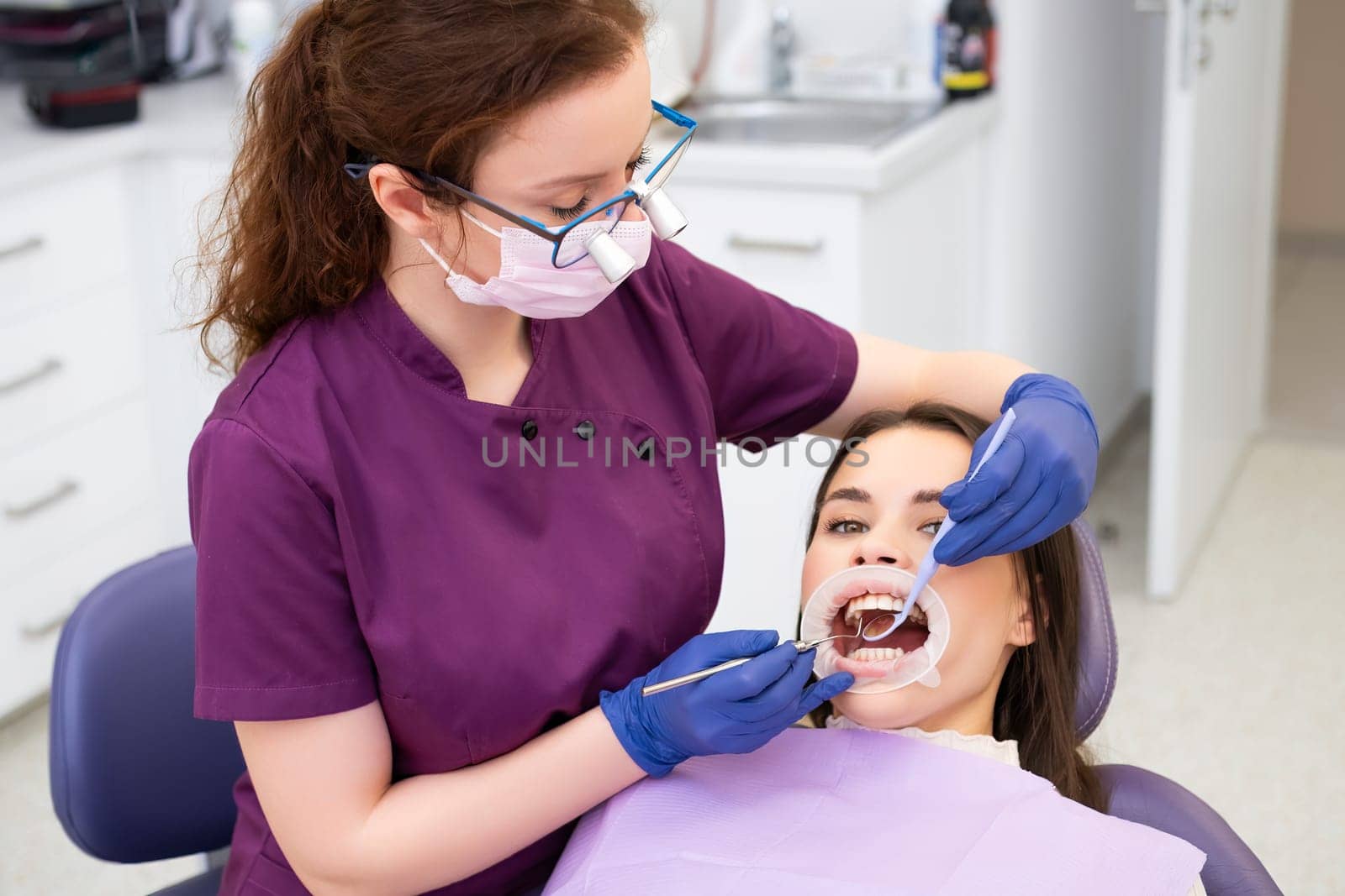 Red haired dentist in glasses performs examination of teeth of brunette female patient by vladimka