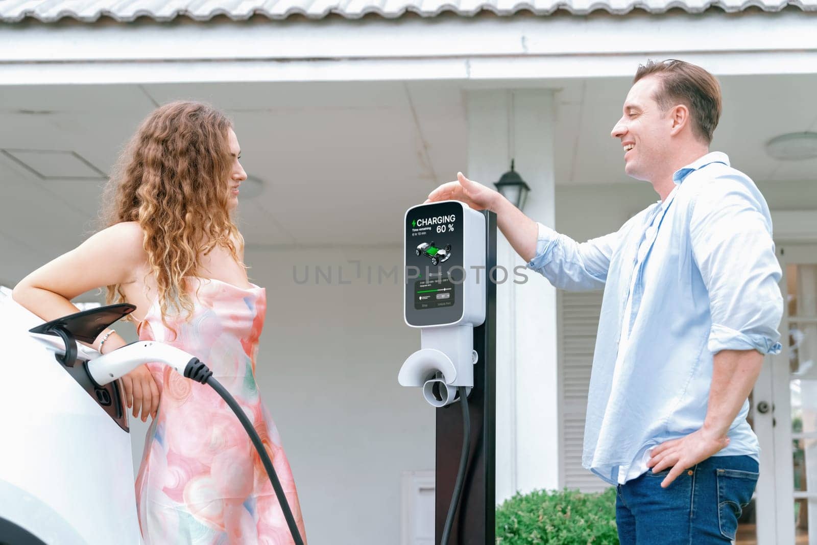 Happy and lovely couple with eco-friendly conscious recharging electric vehicle from EV home charging station. EV car technology utilized for residential home to future sustainability. Synchronos