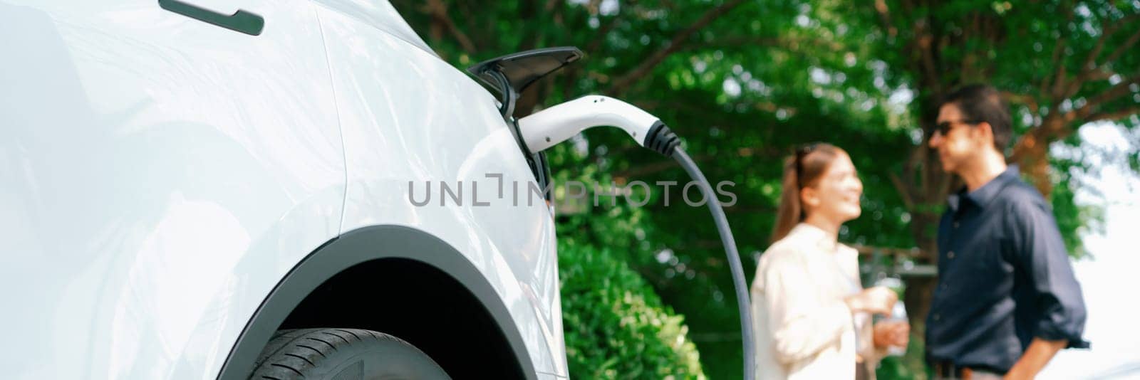 Young couple recharge electric car battery from charging station in green city park in springtime. Rechargeable EV car for sustainable environmental friendly urban travel lifestyle. Panorama Expedient