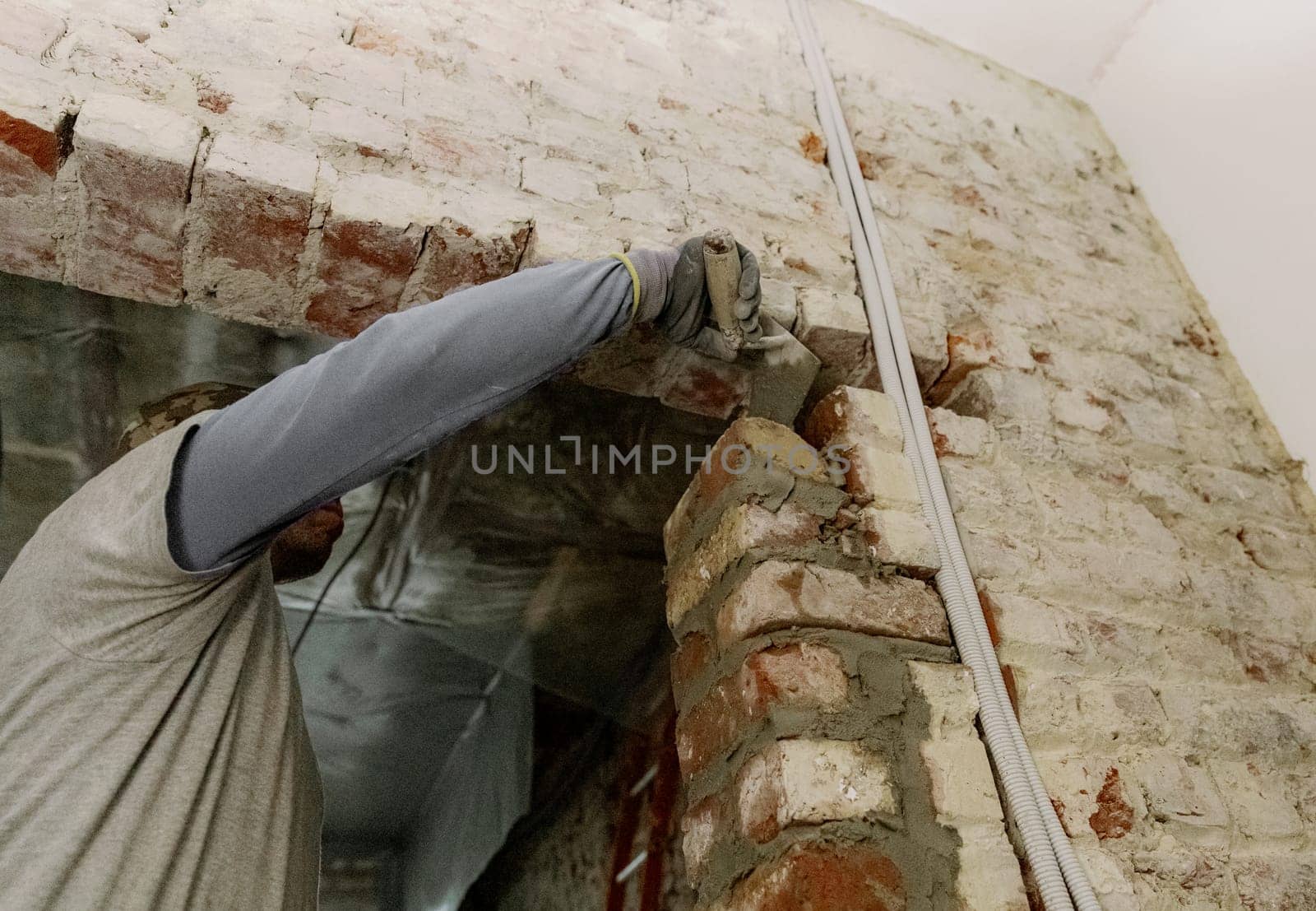 One young Caucasian man puts fresh cement on bricks with a spatula, standing on the right on a stepladder in an old house, close-up view from below.