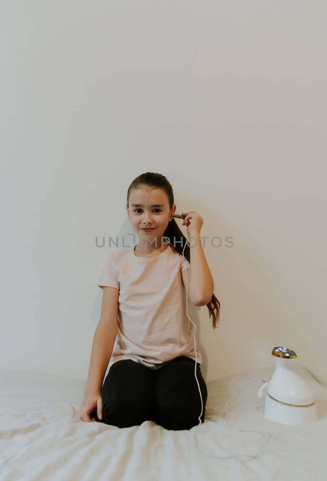 One beautiful Caucasian brunette girl with collected hair and in a pink T-shirt treats her ear with an infrared light device, sitting on her knees on a bed at home near a white wall, close-up side view.