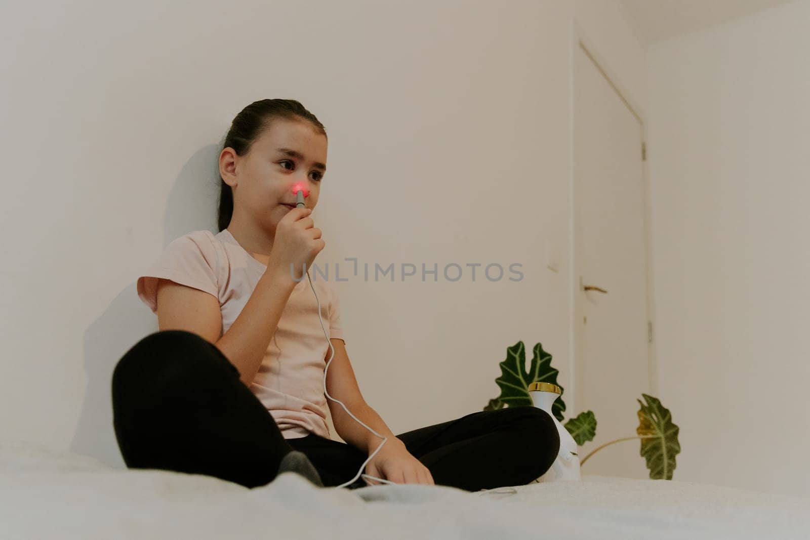 One beautiful Caucasian brunette girl with collected hair and in a pink T-shirt treats the right nasal passage with an apparatus with infrared light, sitting on the bed, cross-legged, leaning on a white wall, close-up view from below.