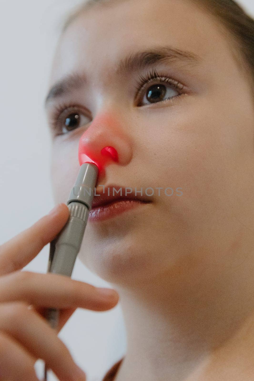 One beautiful Caucasian brunette girl with collected hair and in a pink T-shirt treats the right nasal passage with an apparatus with infrared light, sitting on a bed against a white wall, a very close-up view from below.