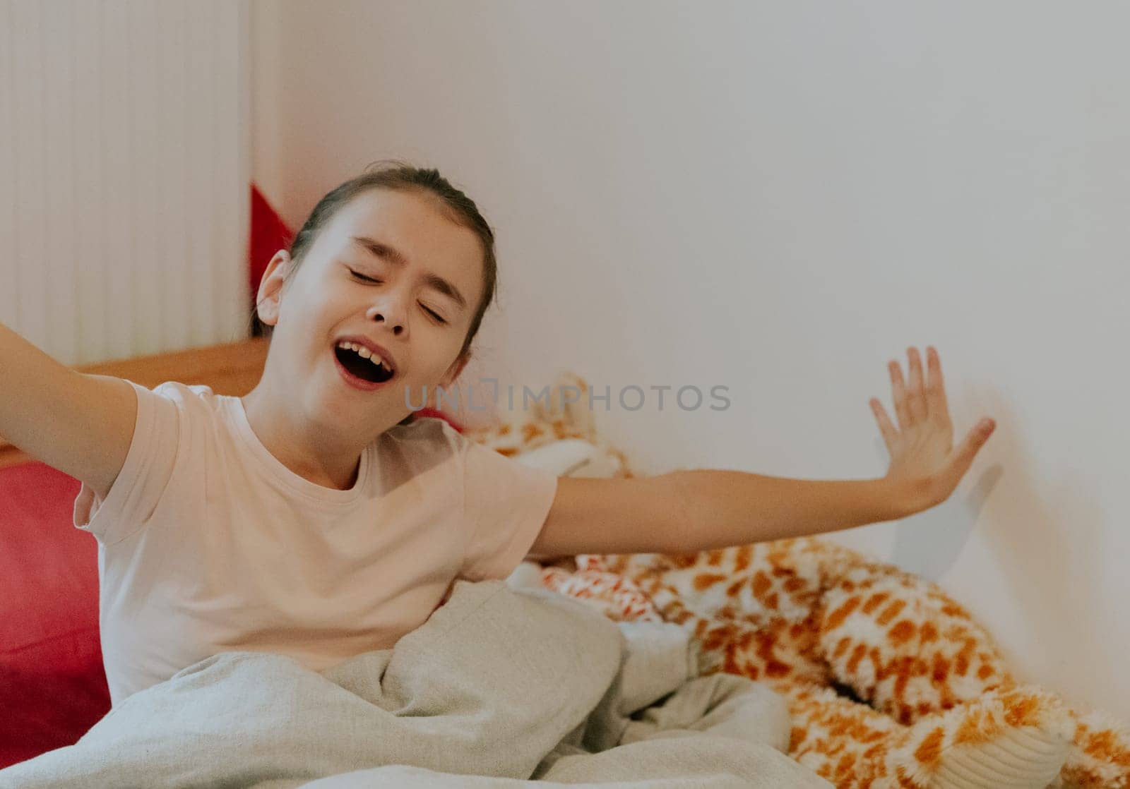 One beautiful little Caucasian brunette girl, waking up, yawns and sits up in bed with her eyes closed, stretches her arms out to the sides, close-up side view.