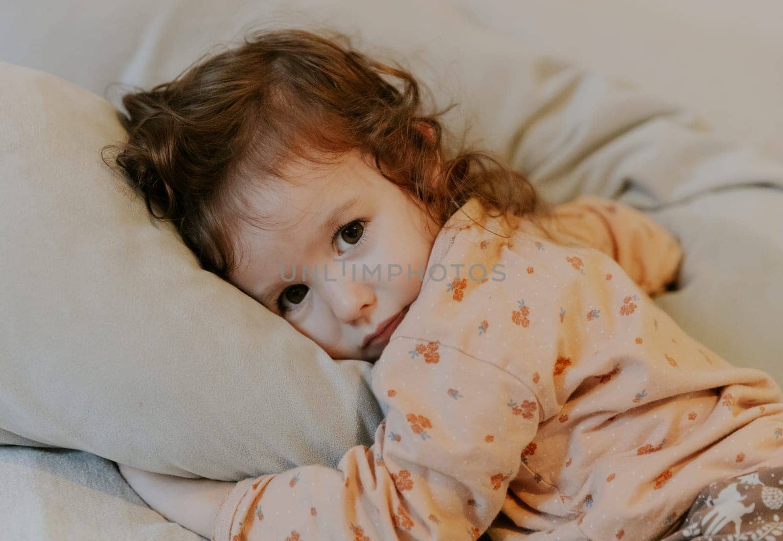 One beautiful little Caucasian girl in pajamas lies on her stomach on the bed hugging a gray pillow early in the morning in the room and looks at the camera, close-up side view.