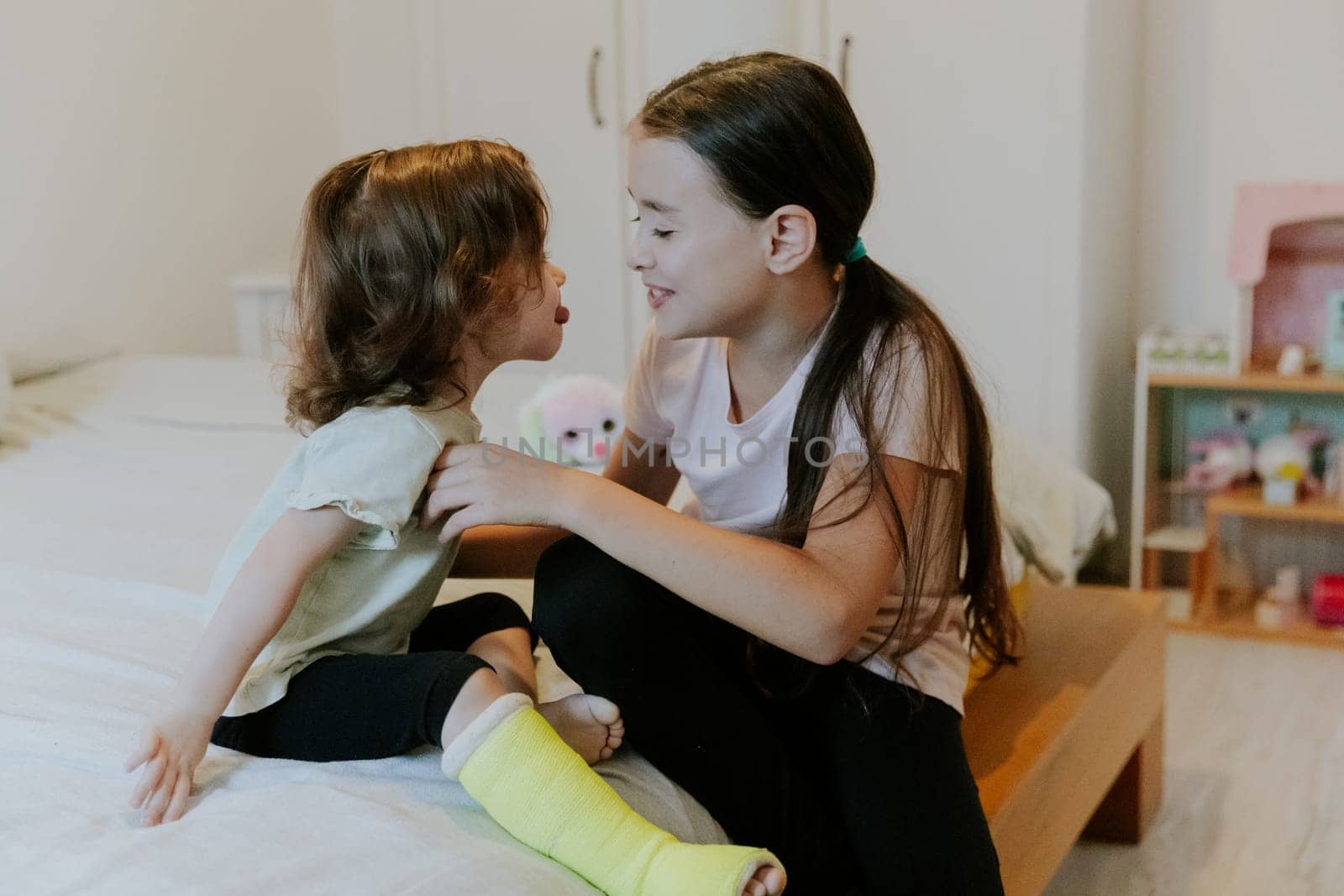 Two beautiful little Caucasian girl with a happy smile on her face fooling around showing her tongue out to each other while sitting on the bed early in the morning in the room, close-up side view with depth of field.