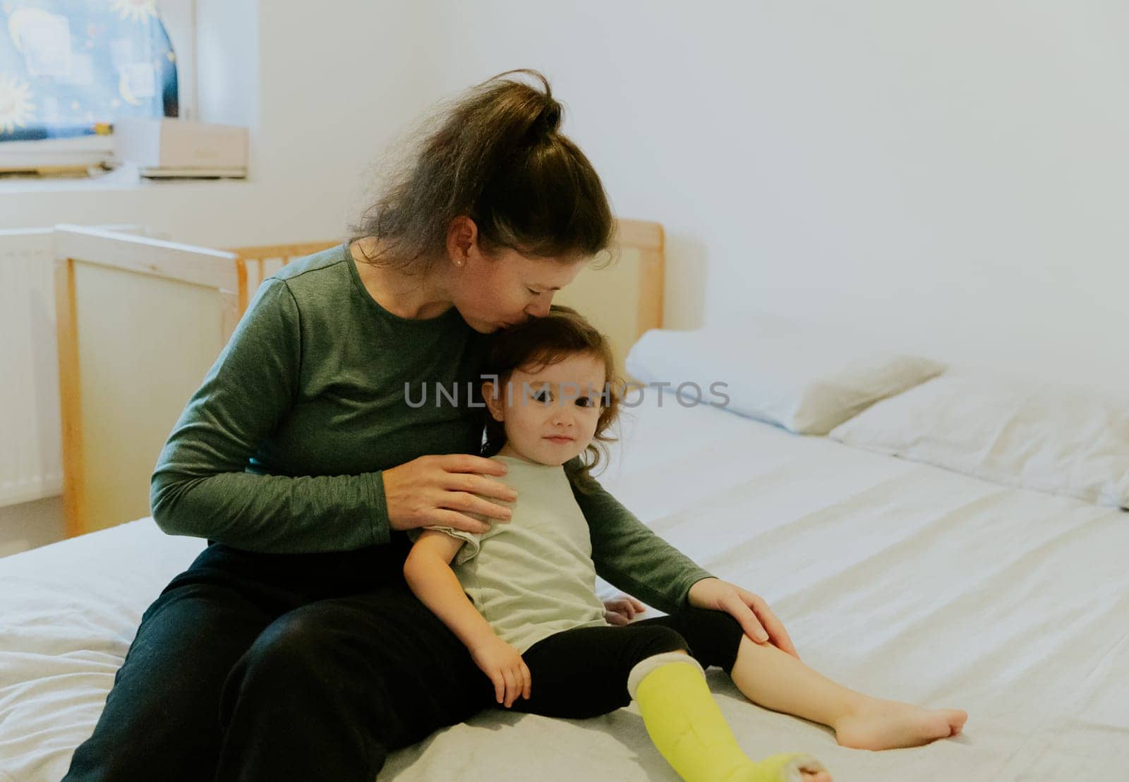 One young girl with a smile on her face gently kisses the head of a little girl with a green cast on her leg, sitting on the bed early in the morning in the room, close-up side view with depth of field.