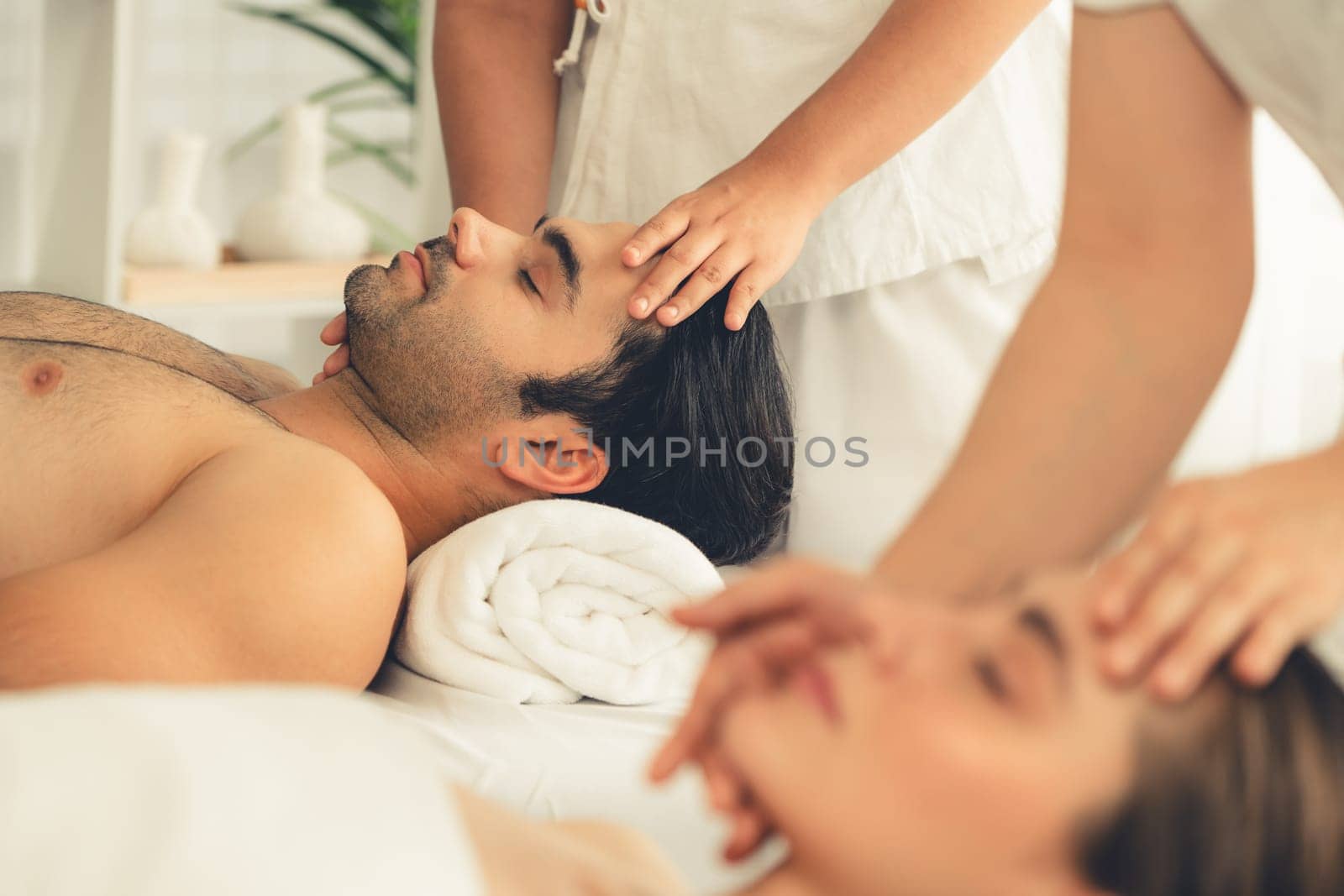 Caucasian couple enjoying relaxing anti-stress head massage and pampering facial beauty skin recreation leisure in dayspa modern light ambient at luxury resort or hotel spa salon. Quiescent