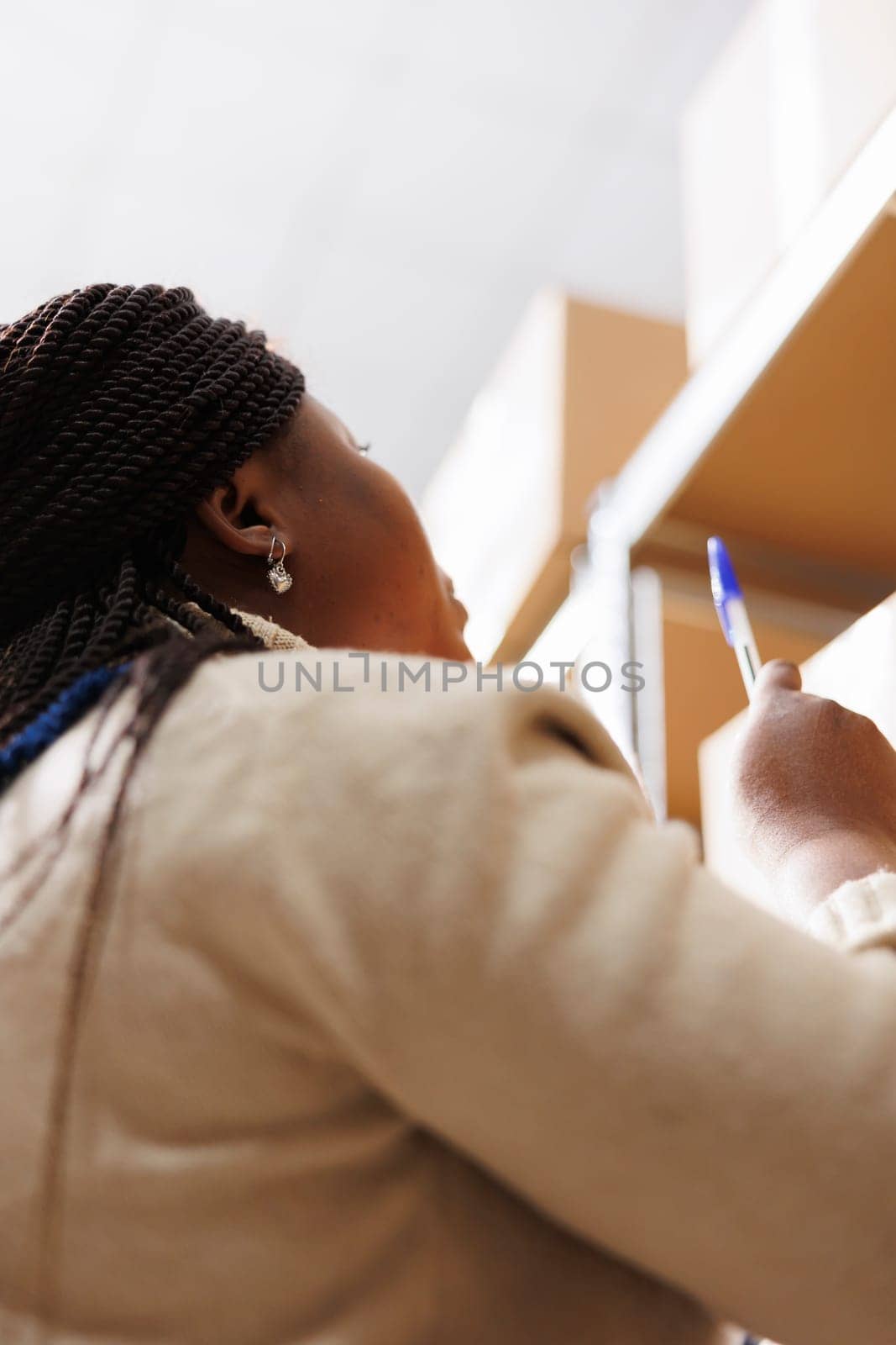 Storehouse worker standing near parcel shelf and pointing upwards with pen. Industrial warehouse african american manager supervising goods storage and dispatching and taking notes