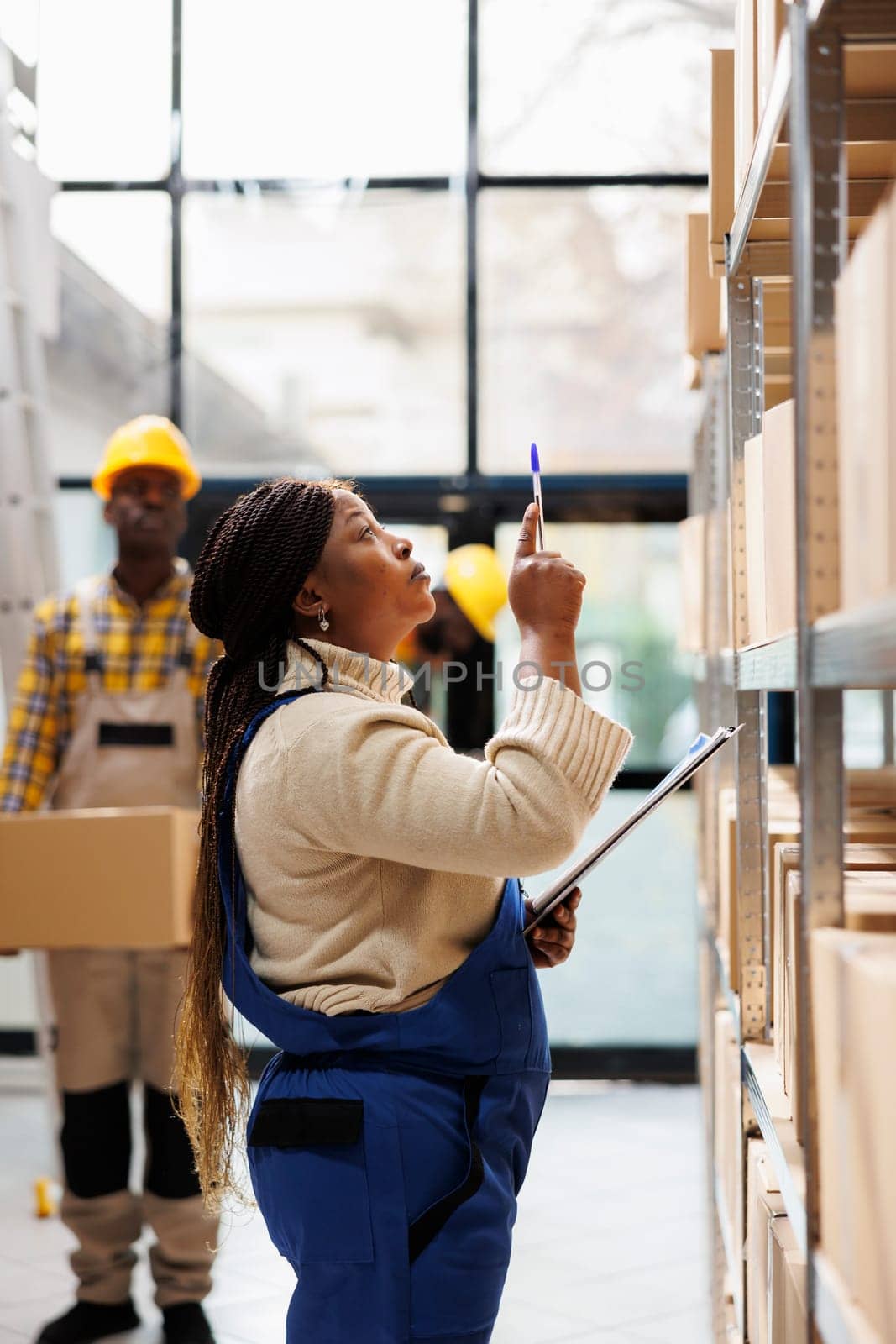 African american woman warehouse manager looking at parcels and controlling goods stock. Logistics operator coordinating products storage and taking notes near cardboard boxes shelf