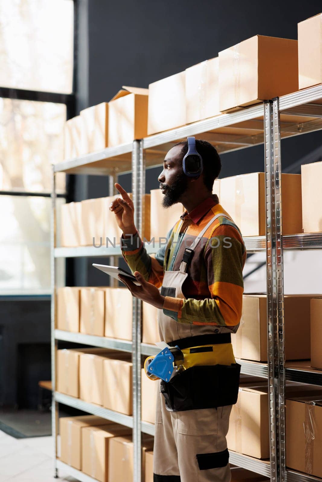 Delivery service manager in headphones searching parcel and counting cardboard boxes on shelf using inventory app. Shipment operator standing in warehouse aisle, listening to music and holding tablet