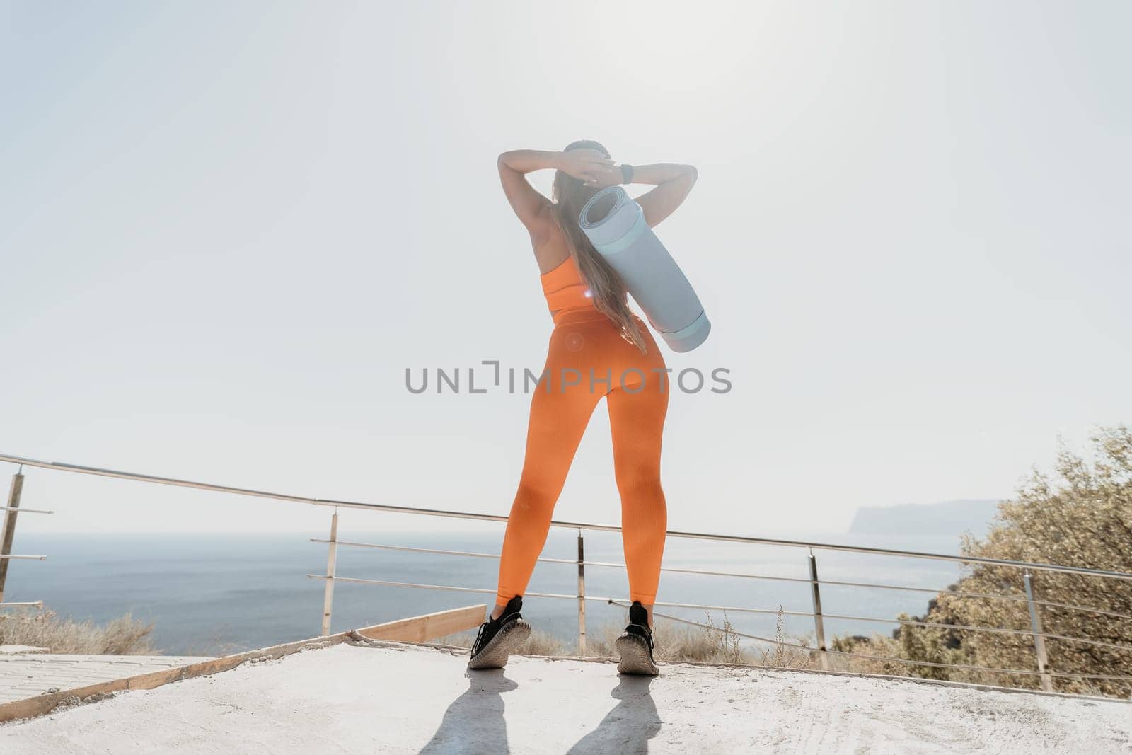 Fitness woman sea. Happy middle aged woman in orange sportswear exercises morning outdoors on yoga mat with laptop in park over ocean beach. Female fitness pilates yoga routine. Healthy lifestyle. by panophotograph