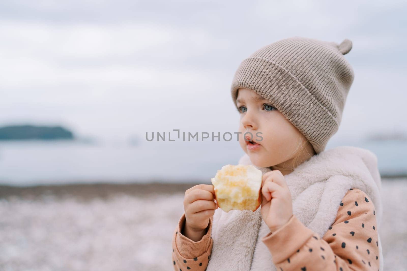 Little girl with a bitten apple in her hands stands by the sea and looks into the distance. High quality photo