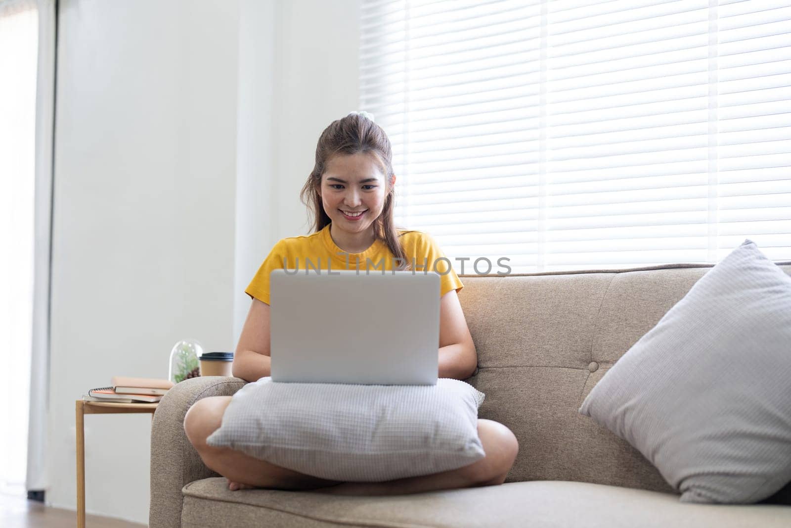 Young woman sitting on the couch and working on project, watching movie on laptop rest and happy chatting with friend in social network at home by nateemee