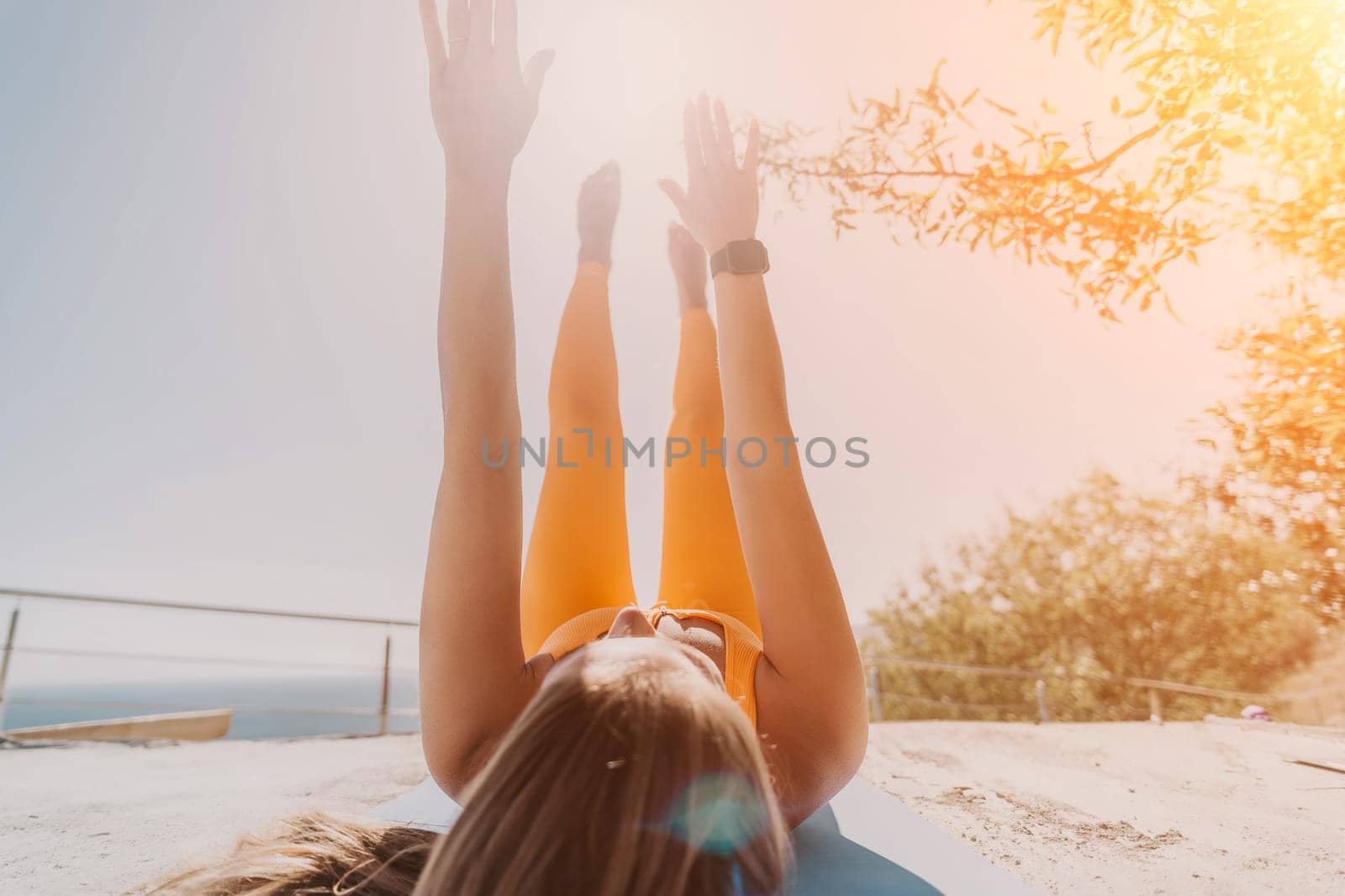 Fitness woman sea. Happy middle aged woman in orange sportswear exercises morning outdoors on yoga mat with laptop in park over ocean beach. Female fitness pilates yoga routine. Healthy lifestyle. by panophotograph