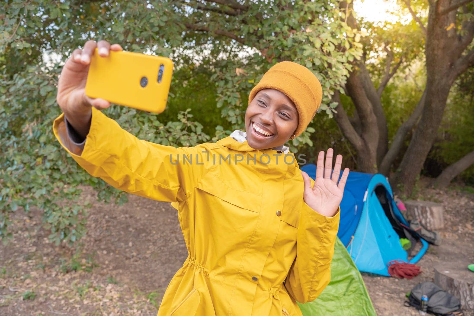 A happy woman wearing a yellow raincoat is smiling as she takes a selfie with her cell phone, surrounded by nature. High quality photo