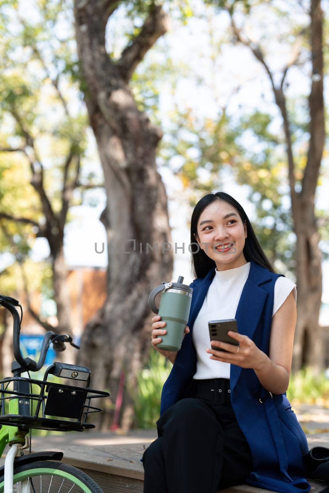 Asian businesswoman with bicycle using smartphone and sitting outside the office building. Woman commuting on bike go to work. Eco friendly vehicle, sustainable lifestyle concept.