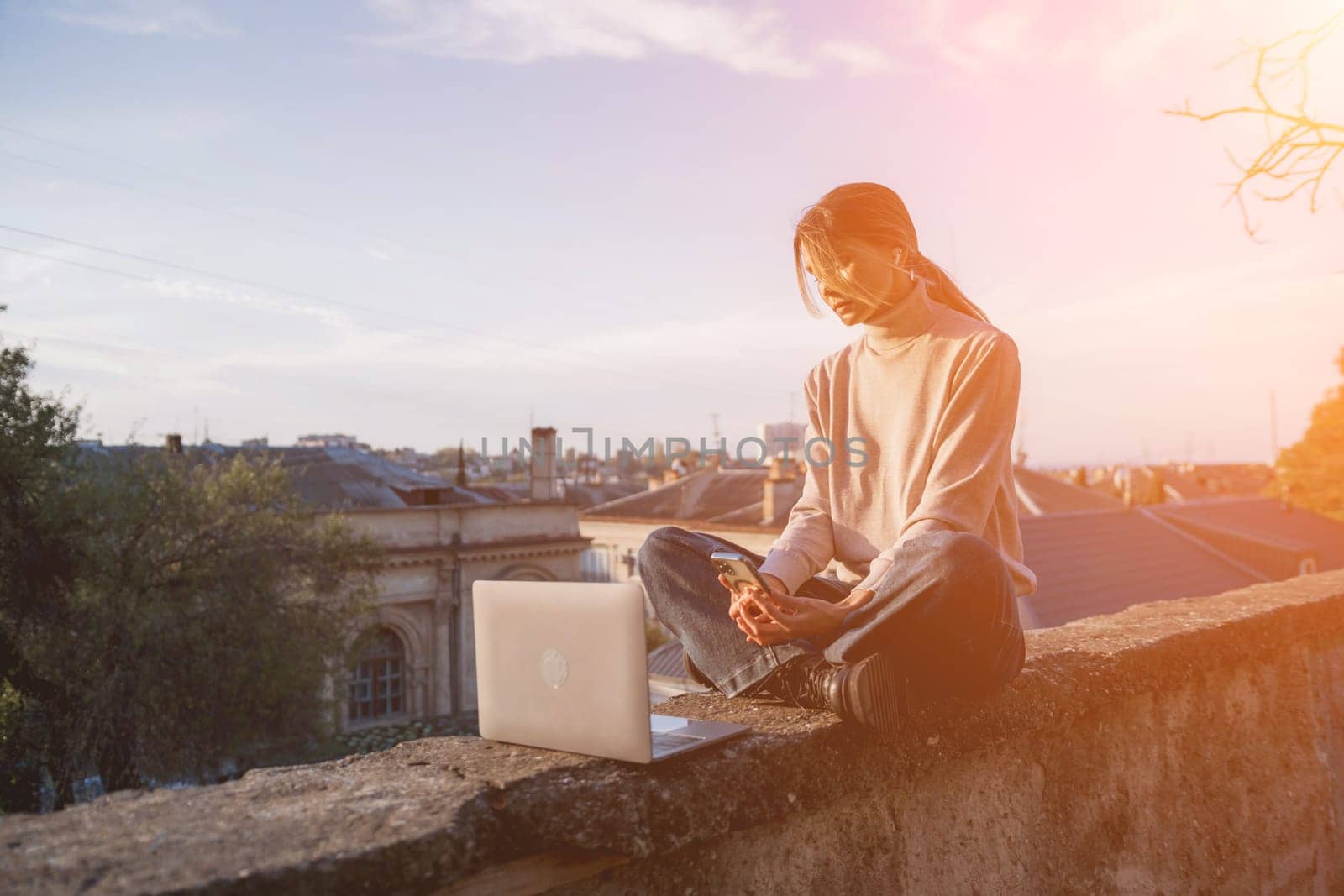 Woman freelancer uses laptop on cement wall outdoors against the sky and the roof of the city. The woman to be focused on her work or enjoying some leisure time while using her laptop. by Matiunina