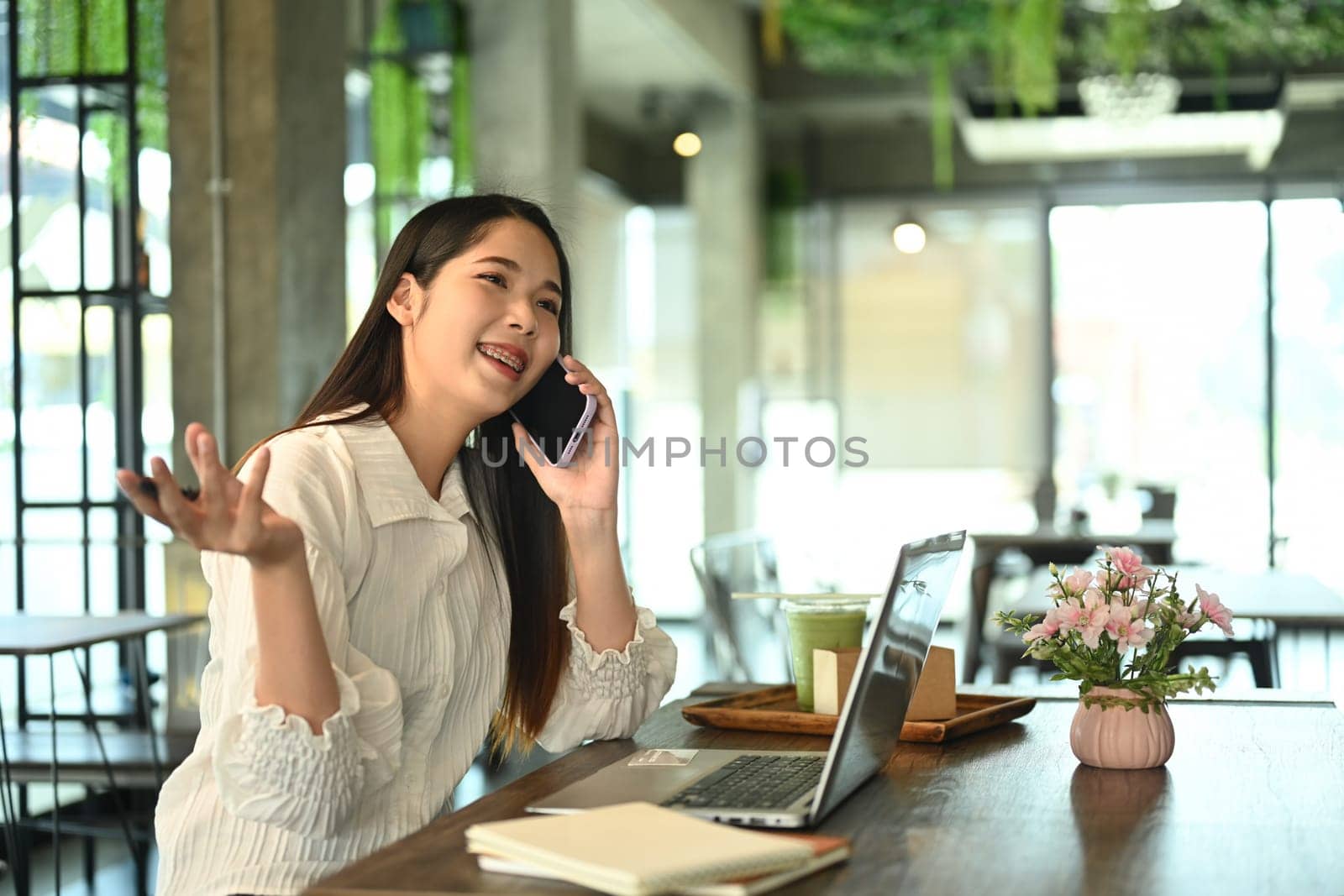 Cheerful young businesswoman having phone conversation and using laptop at cafe by prathanchorruangsak