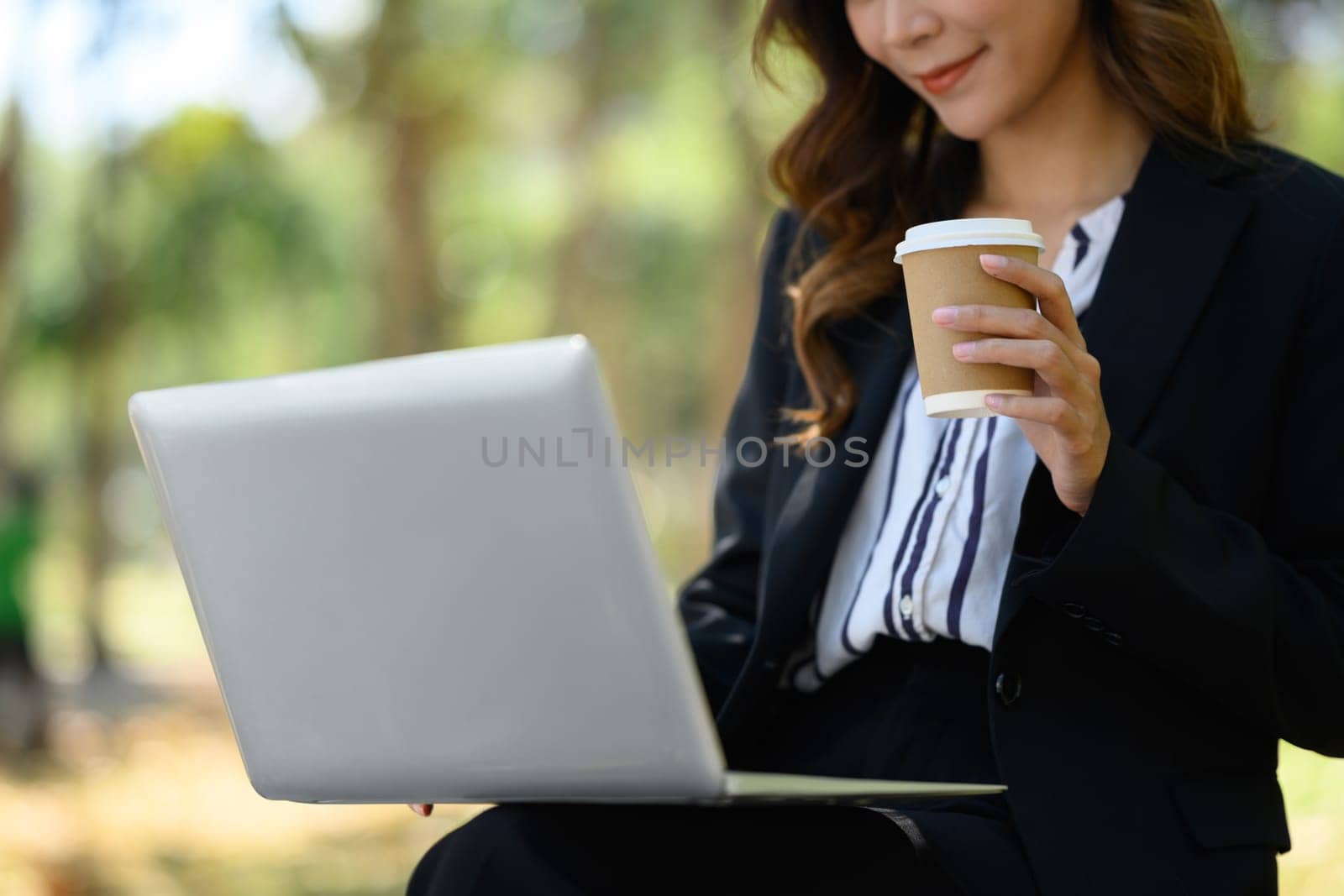 Smiling pretty woman in business suit holding paper cup and using laptop in the city park by prathanchorruangsak