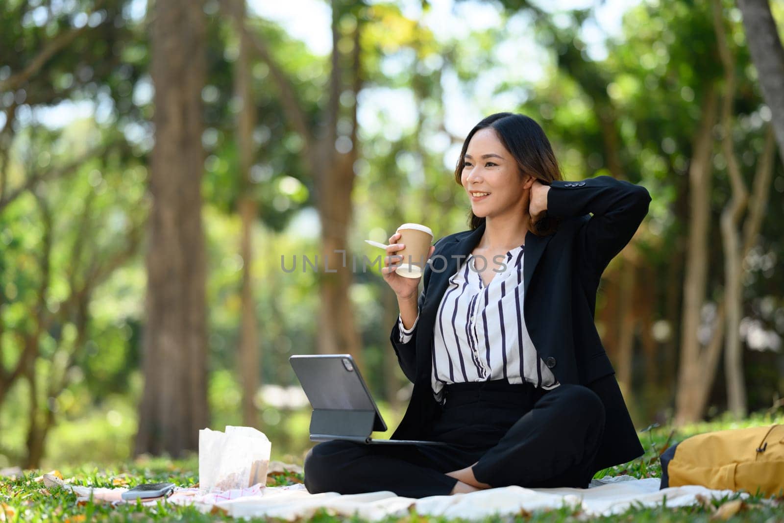 Pleased businesswoman drinking coffee and using digital tablet in the park on beautiful day.
