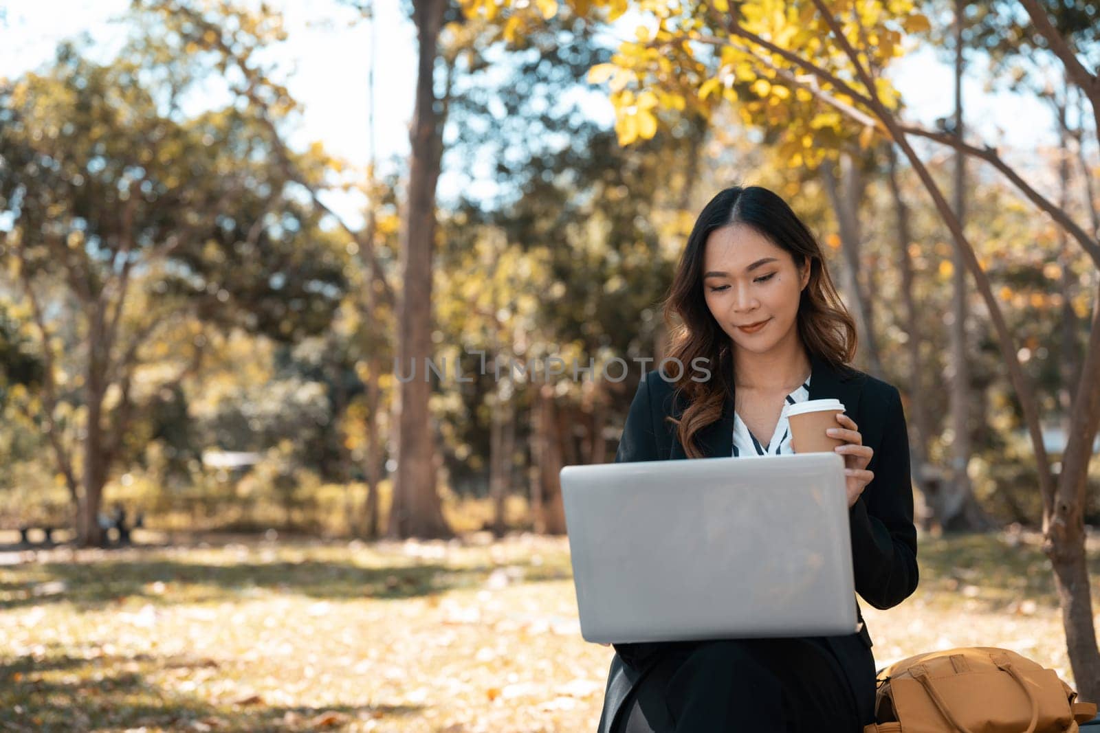 Portrait of young businesswoman with cup of coffee using laptop on bench in autumn park by prathanchorruangsak