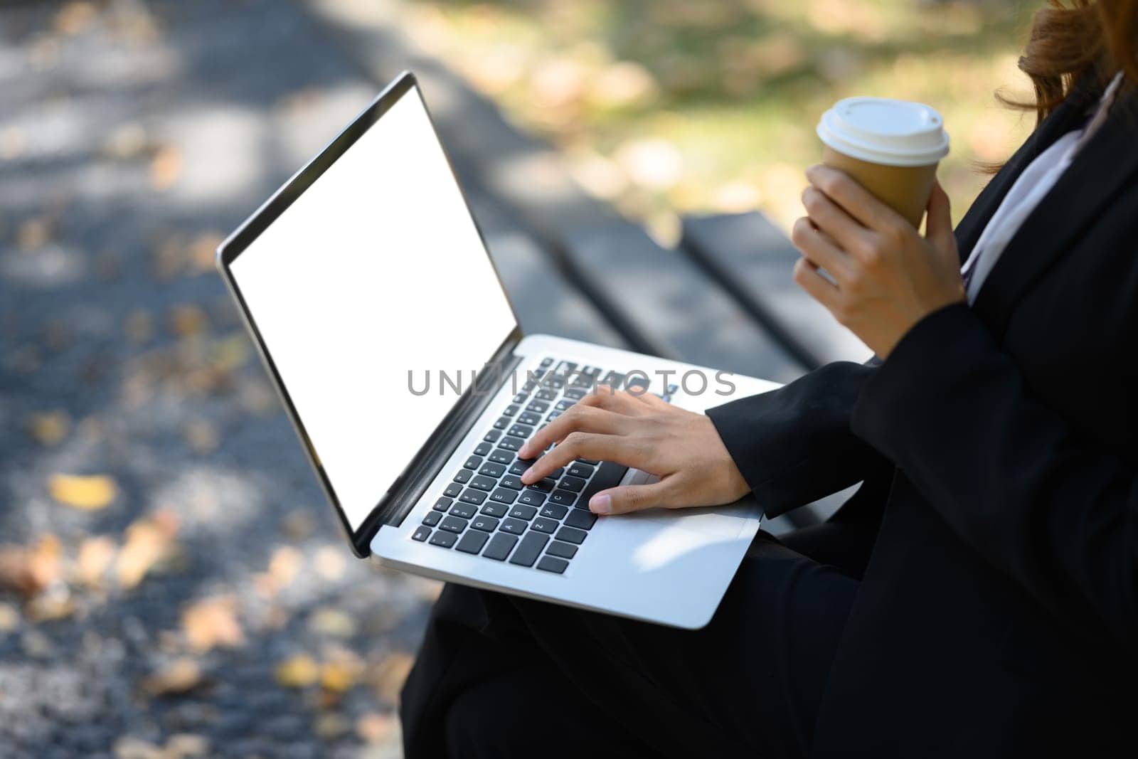 Cropped shot of businesswoman with paper cup of coffee in hand using laptop on a park bench by prathanchorruangsak