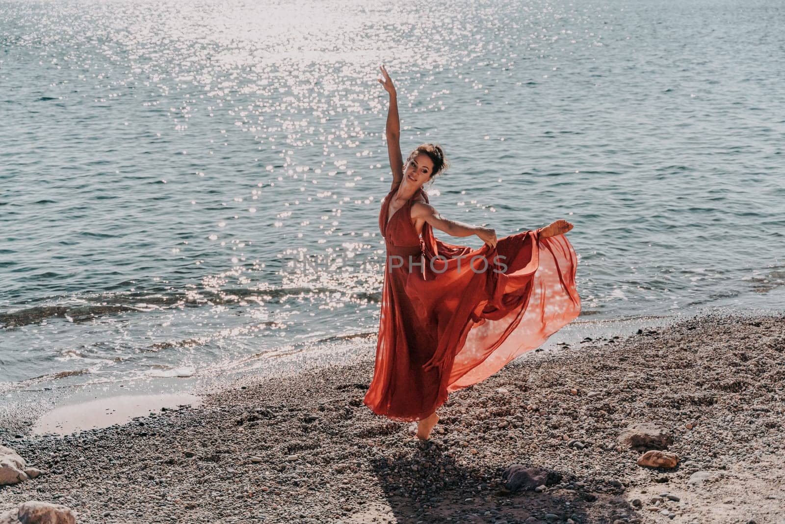 Woman red dress sea. Female dancer in a long red dress posing on a beach with rocks on sunny day. Girl on the nature on blue sky background