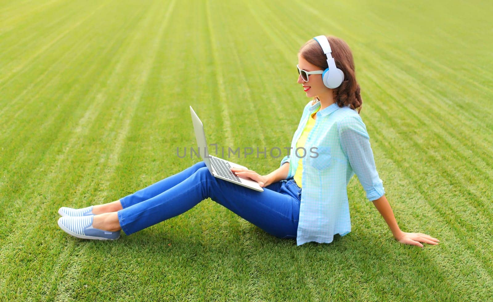 Happy young woman with laptop listening to music in headphones in the park on green grass background