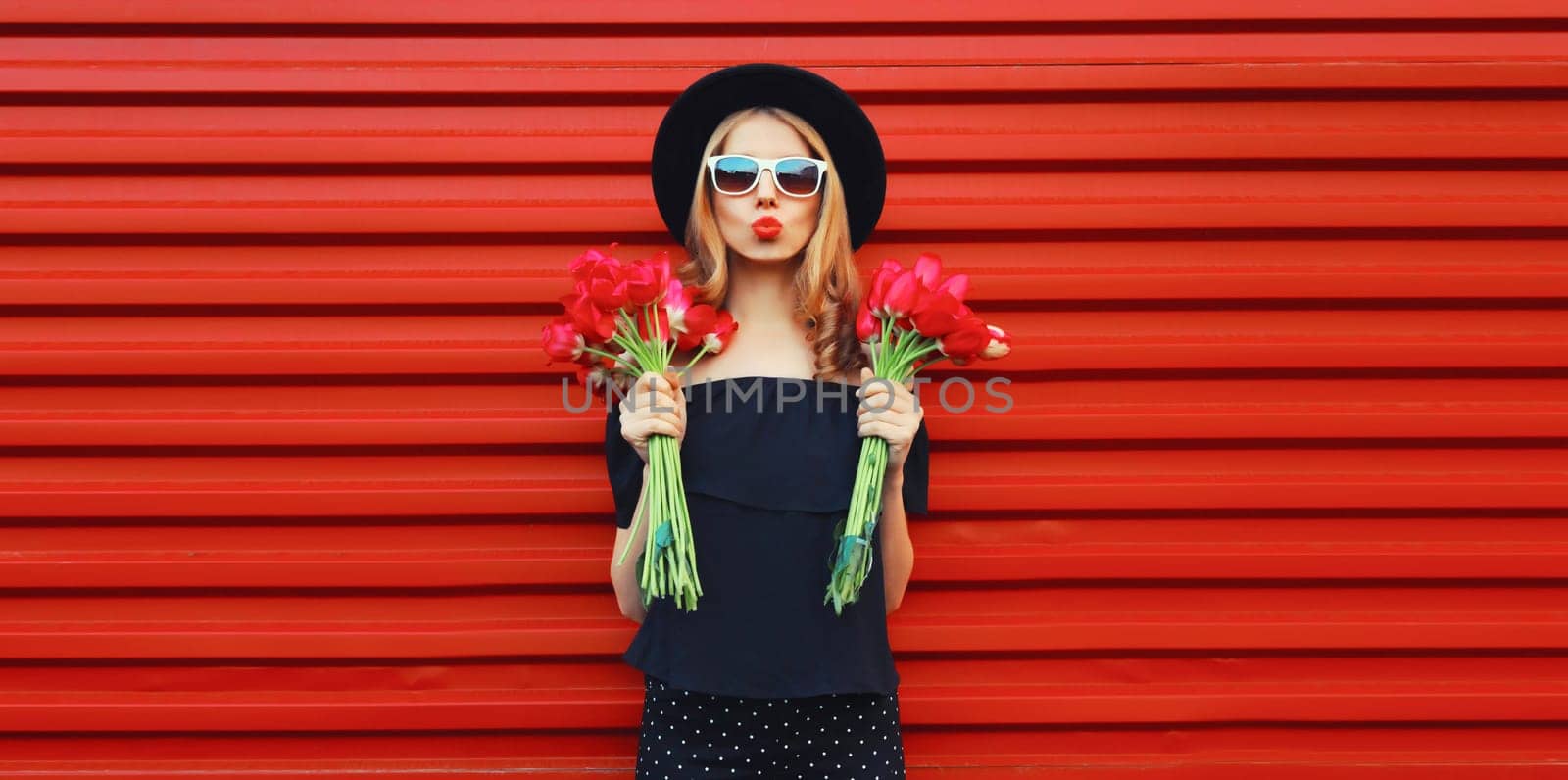 Beautiful stylish woman with bouquet of red rose flowers blowing kiss in black round hat by Rohappy