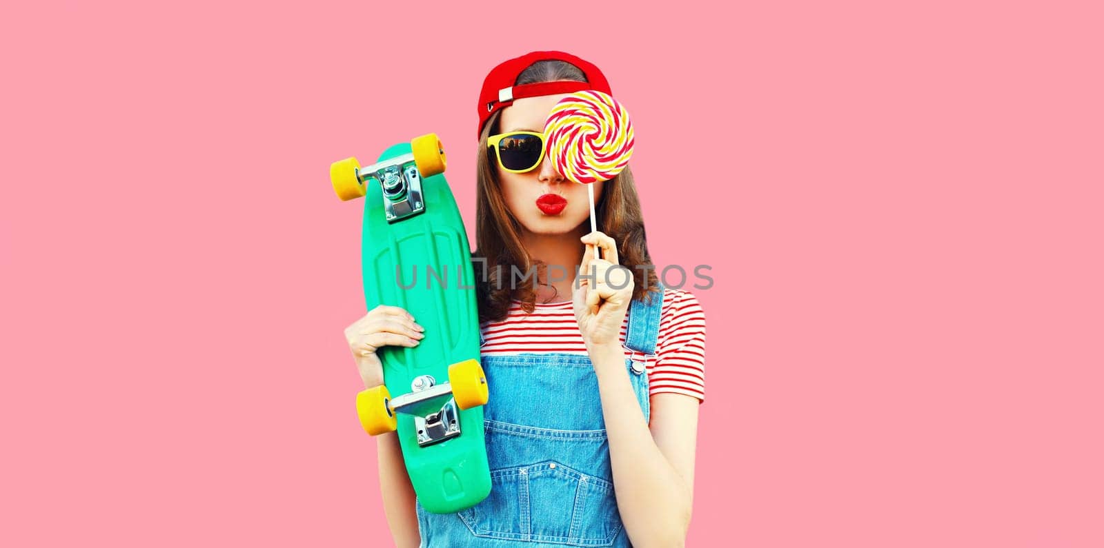 Summer portrait of young woman with lollipop and green skateboard on pink background