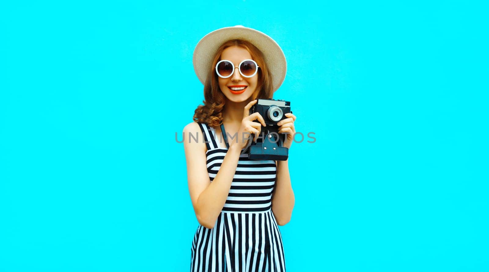 Summer portrait of happy smiling young woman photographer with vintage film camera wearing straw hat, dress, sunglasses on blue background