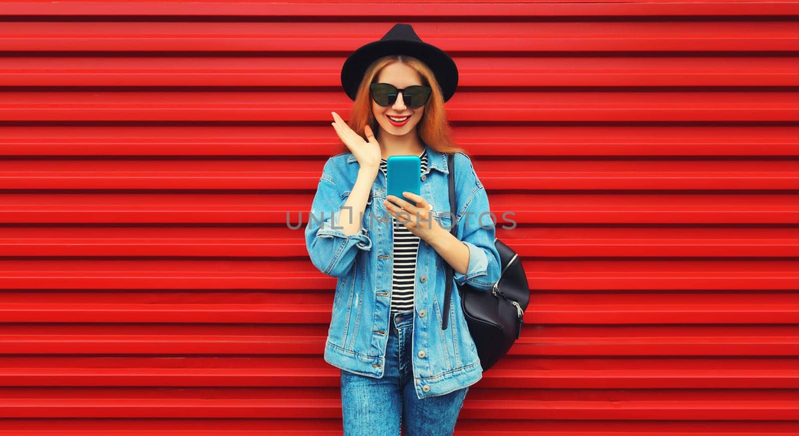 Portrait of stylish modern smiling young woman with smartphone wearing jean jacket, black round hat on red background