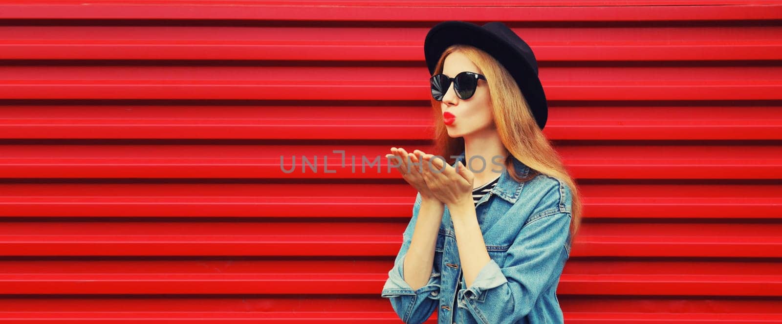 Stylish young woman model blowing kiss in jean jacket, black round hat on red background by Rohappy