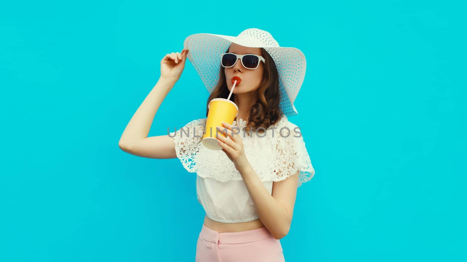 Portrait of beautiful caucasian young woman model with cup of coffee or fresh juice wearing white summer straw hat on studio blue background