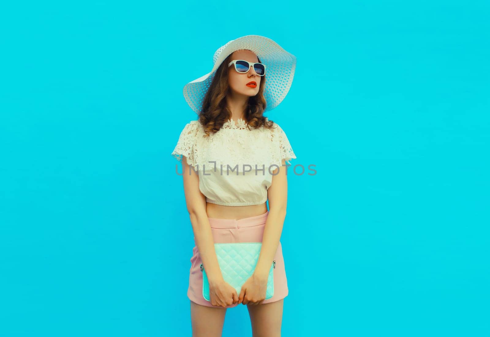 Beautiful caucasian young woman model posing with handbag clutch wearing white summer straw hat on blue studio background