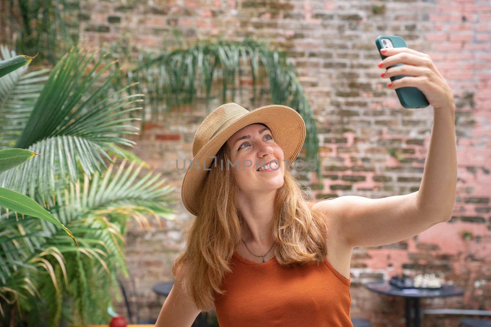 Woman wearing a sun hat is smiling and gesturing while taking a selfie with her cell phone in a leisurely travel landscape surrounded by terrestrial plants. She looks happy in the sunny weather