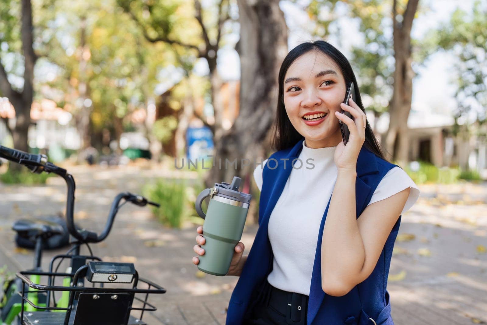 Smiling asian businesswoman hold reusable eco-friendly ecological cup and using mobile while sitting at park by itchaznong
