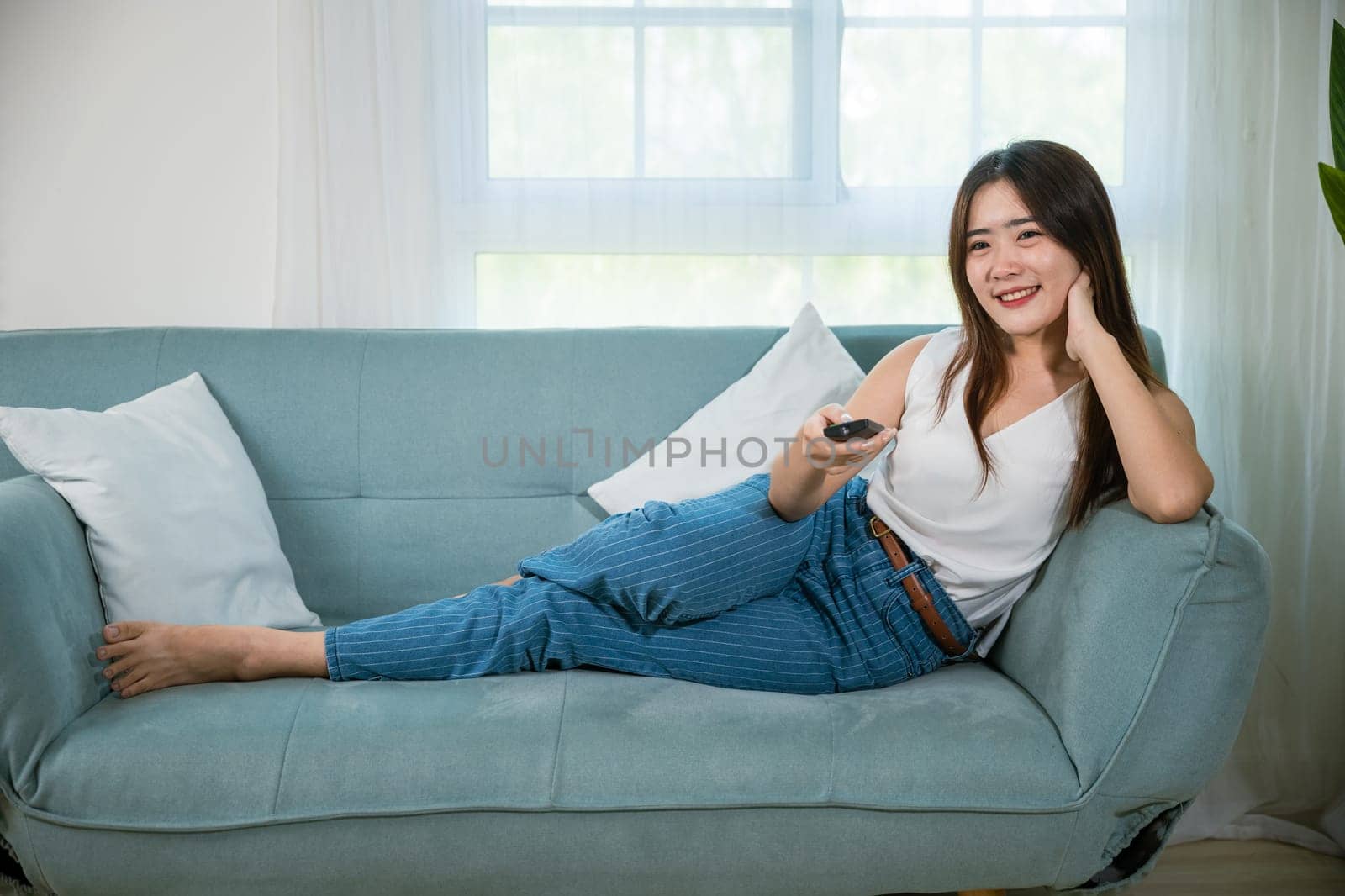 Asian young woman smiling sitting relax watch TV holding remote control on sofa in living room, Happy female fun movie holding remote watching television, Activity lifestyles concept