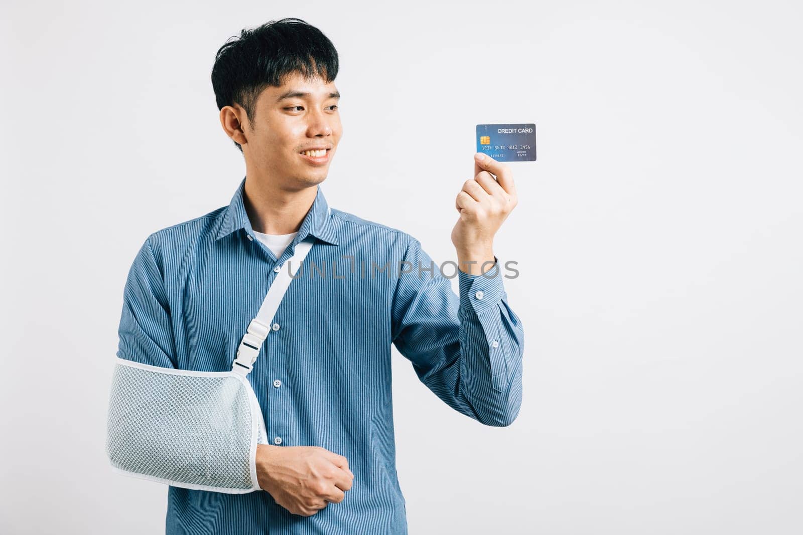 Injured man confidently smiles with a damaged arm in a support splint, managing medical expenses via credit card. Happy Asian man with sling support hand, isolated on white, showcasing health care.