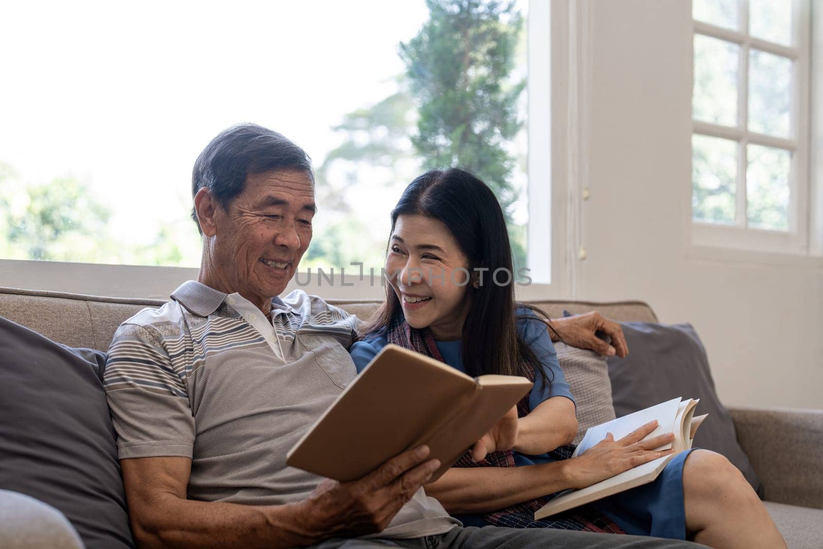 Asian senior couple reading book diary together, sitting on cozy sofa in home interior. Happy retirement activity lifestyle at home by nateemee