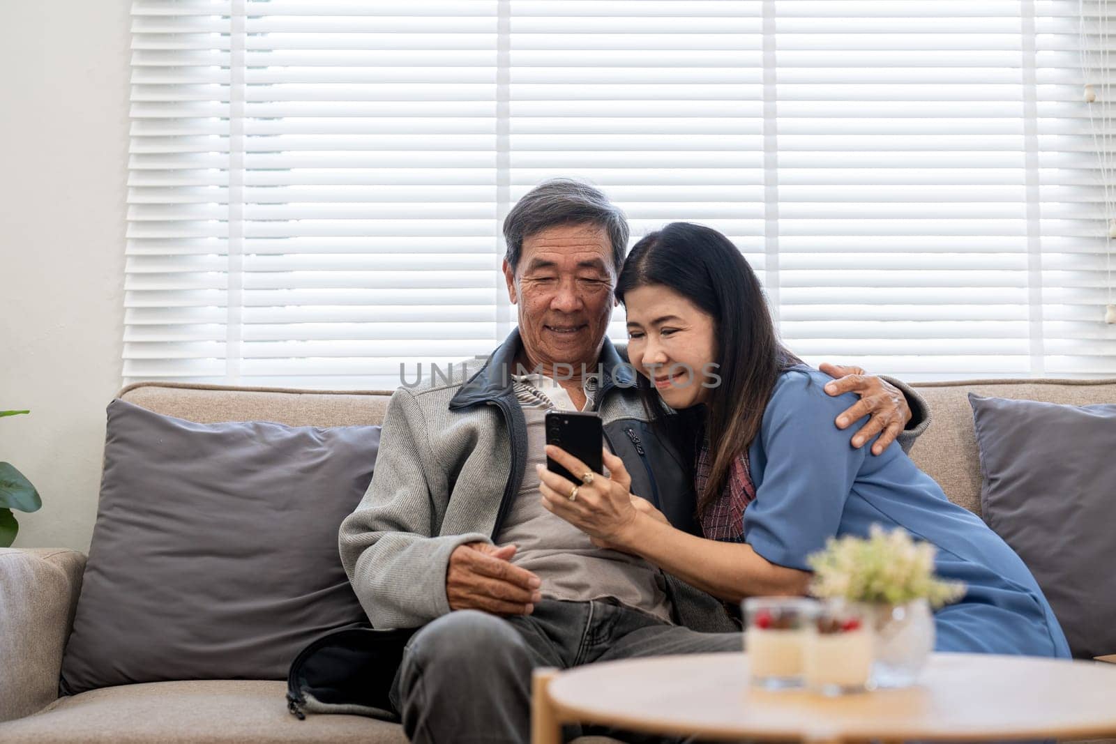 Happy elderly asian couple using smartphone sit on sofa doing ecommerce shopping online on website at home by nateemee