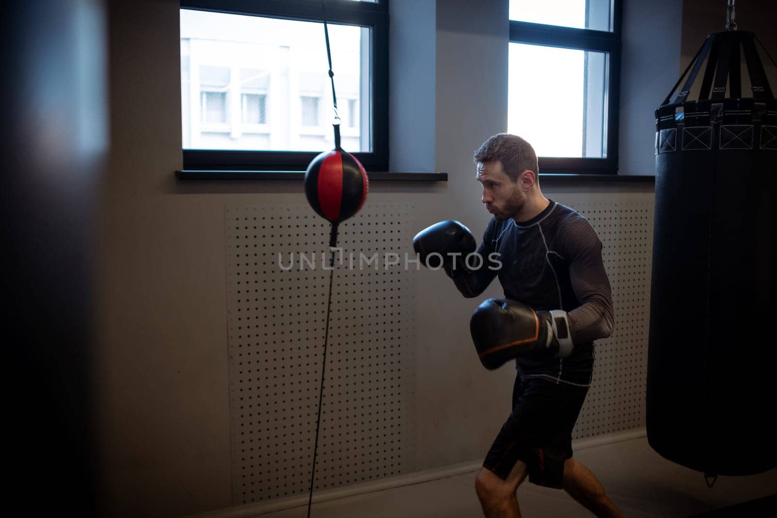 Concentrated young adult bearded boxer honing skills on double-end punching bag, working out focus and accuracy in dimly boxing gym